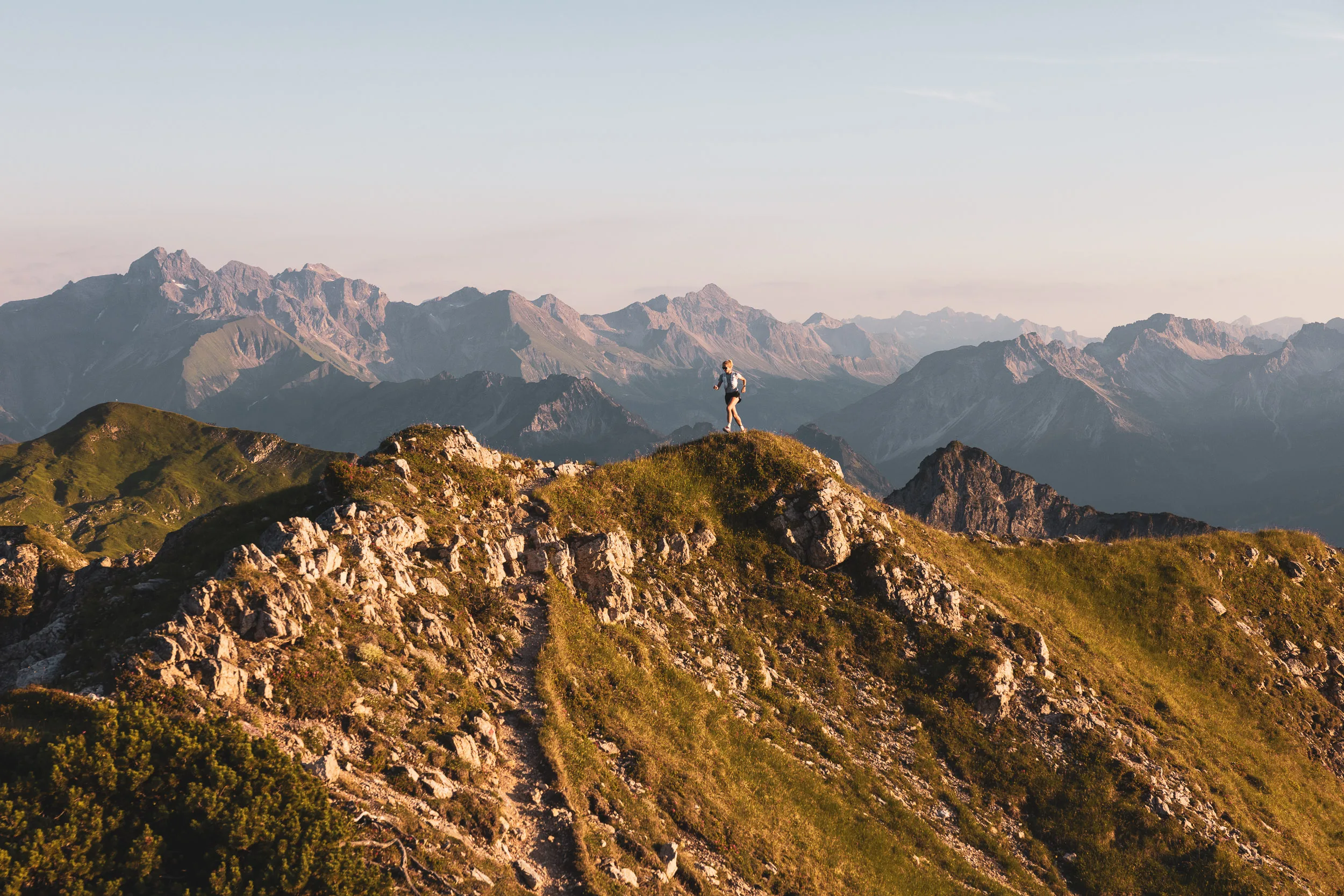 Einzelne Trailrunnerin auf einem weiten Grat vor dem Alpenpanorama
