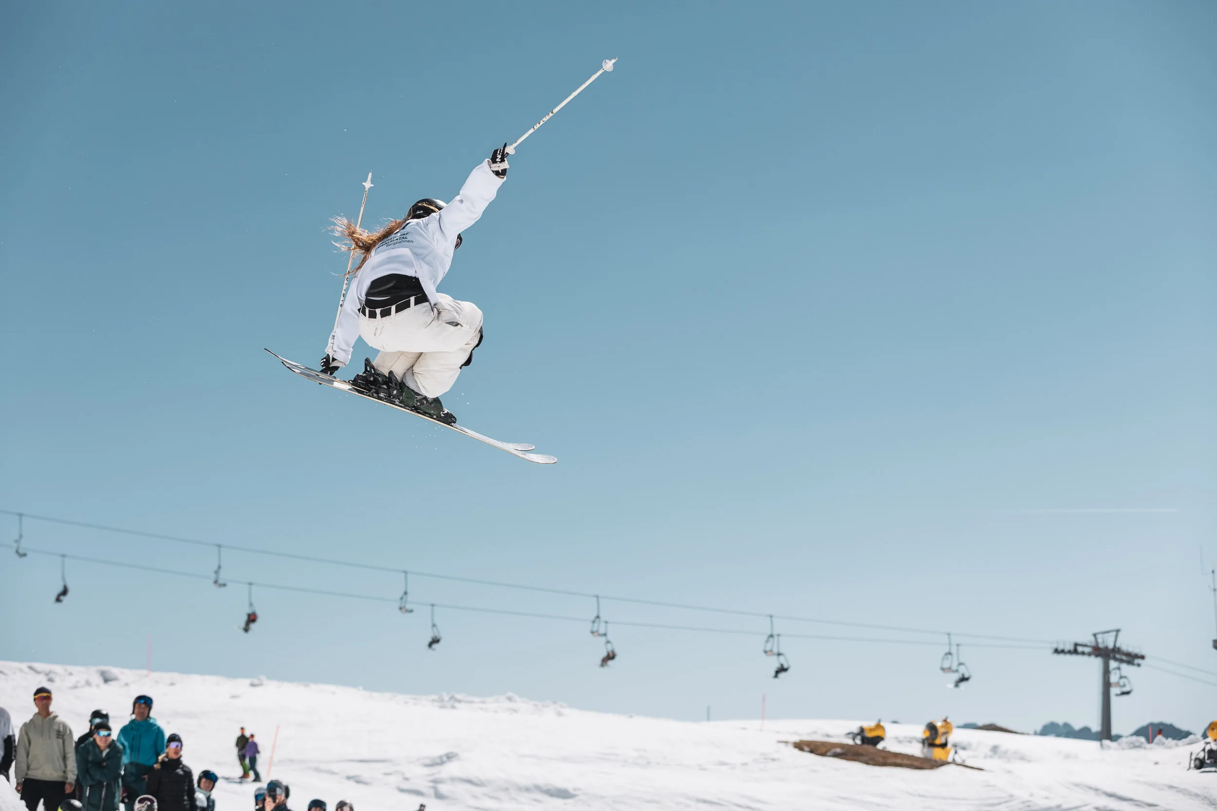 Skifahrer mit Grab hoch in der Luft über einem Kicker vor blauem Himmel, unten das Publikum am Lift