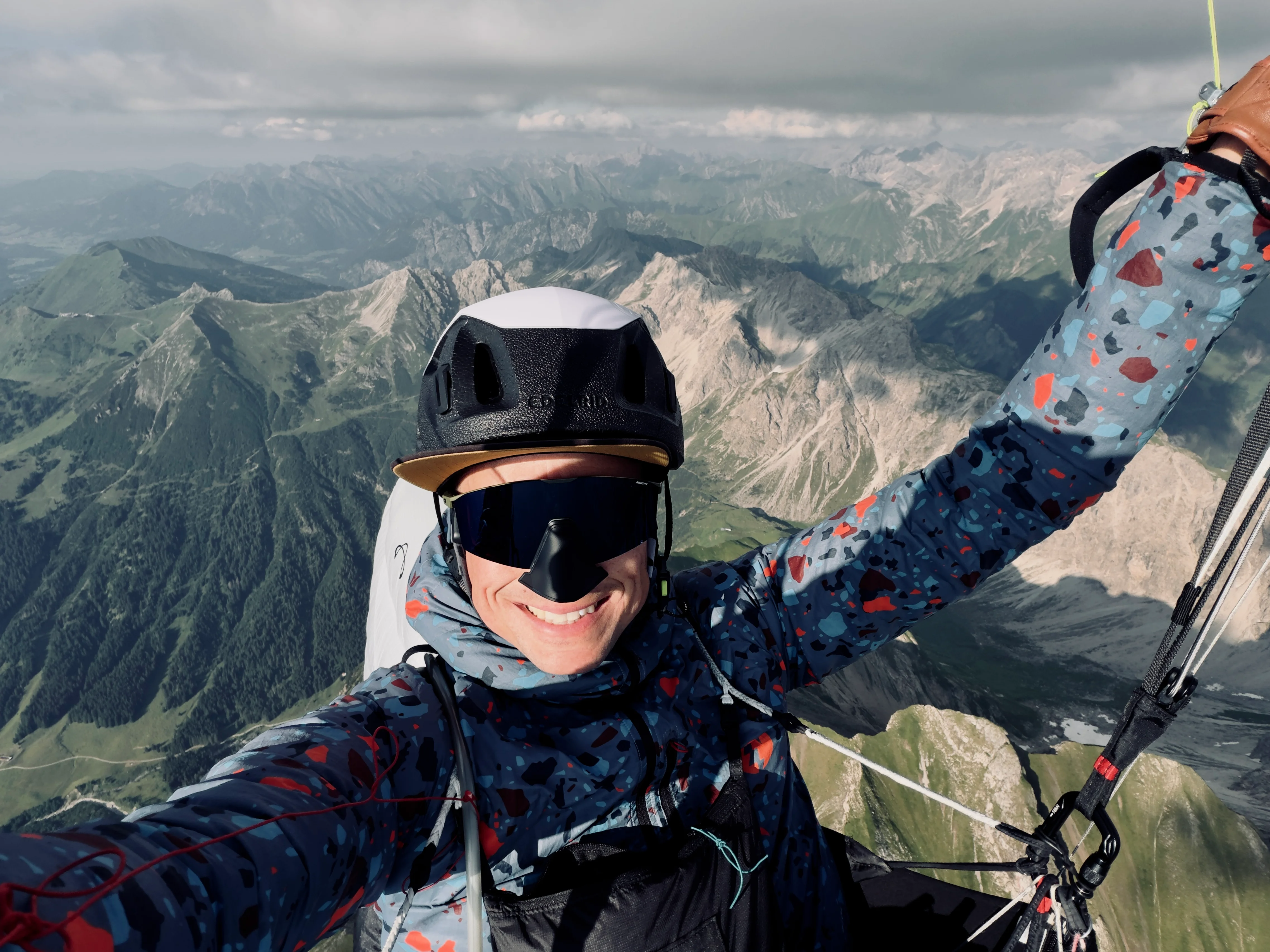 Bastian Morell beim Gleitschirmflug über einer Berglandschaft, mit Helm und Sonnenbrille.