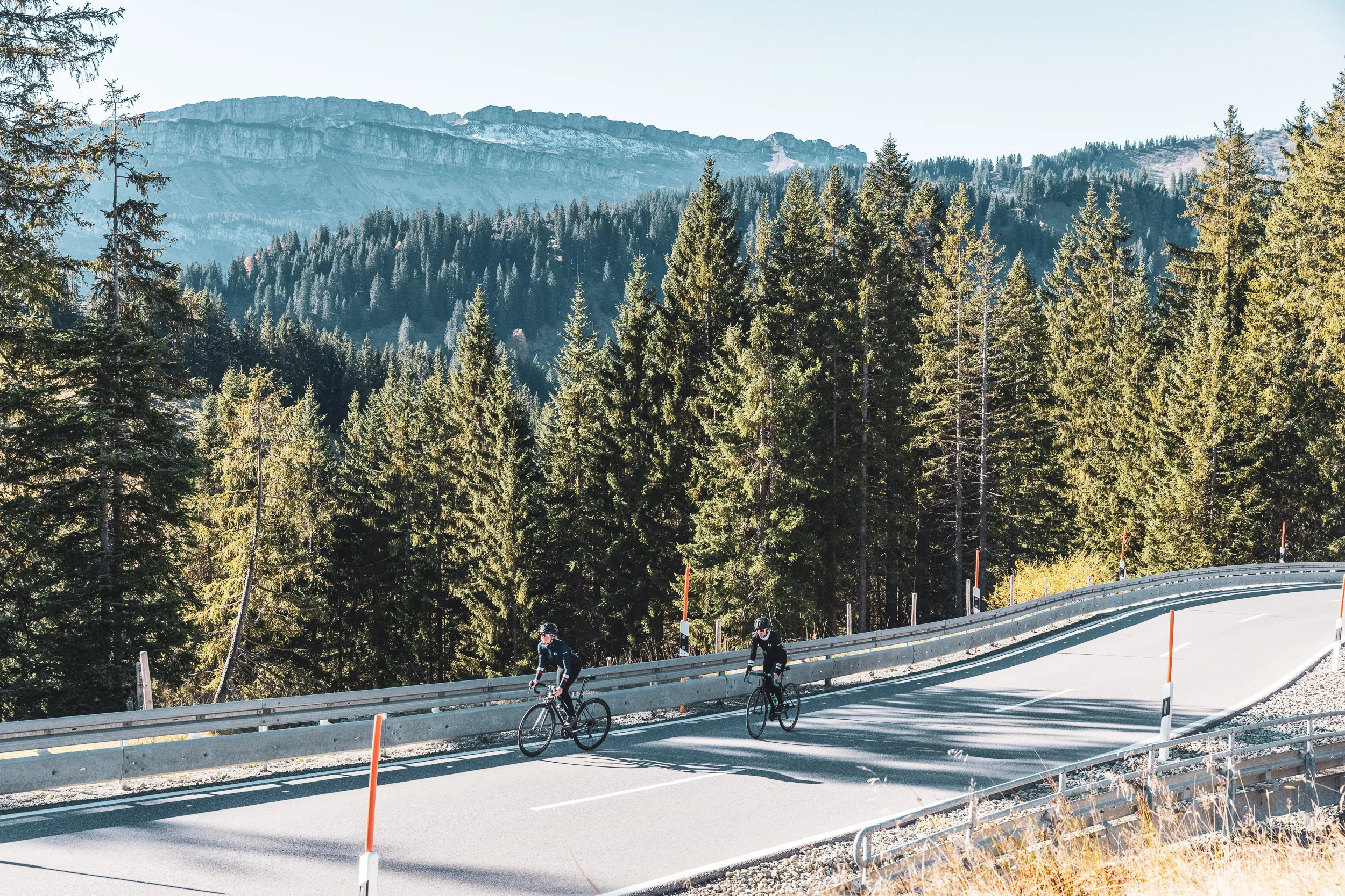 Zwei Rennradfahrerinnen fahren vor Bergwald und Felsen über den Pass