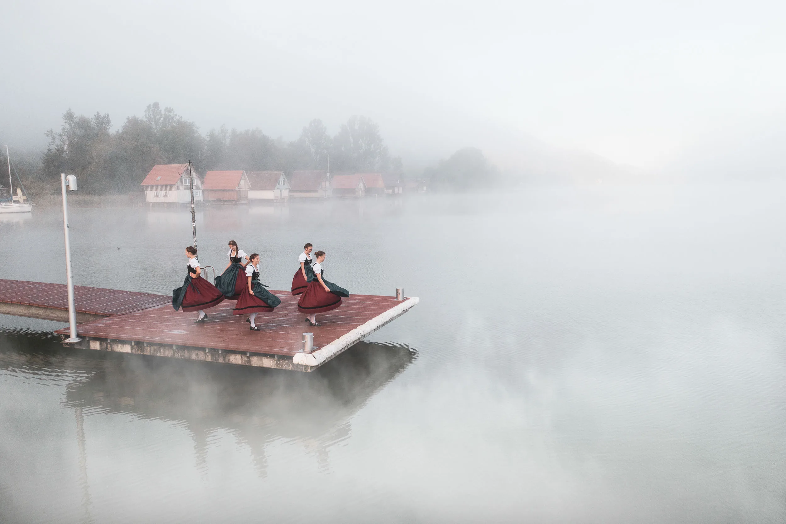 Tänzerinnen in Tracht auf einem Steg am Alpsee, umgeben von Nebel und ruhigem Wasser.