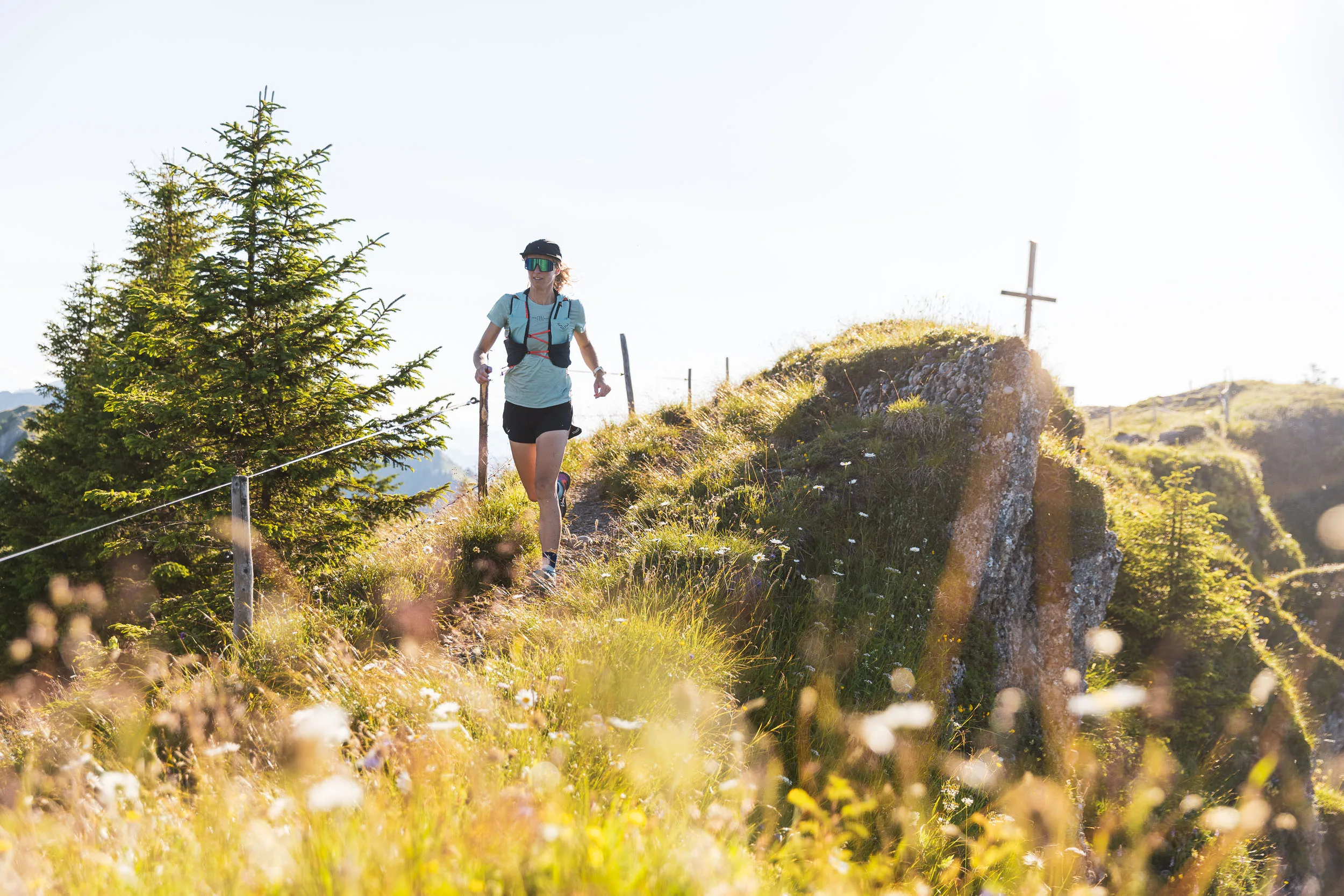 Eine Trailrunnerin läuft zwischen Bergblumen und einer Felskante bergab, hinter ihr steht ein Gipfelkreuz.