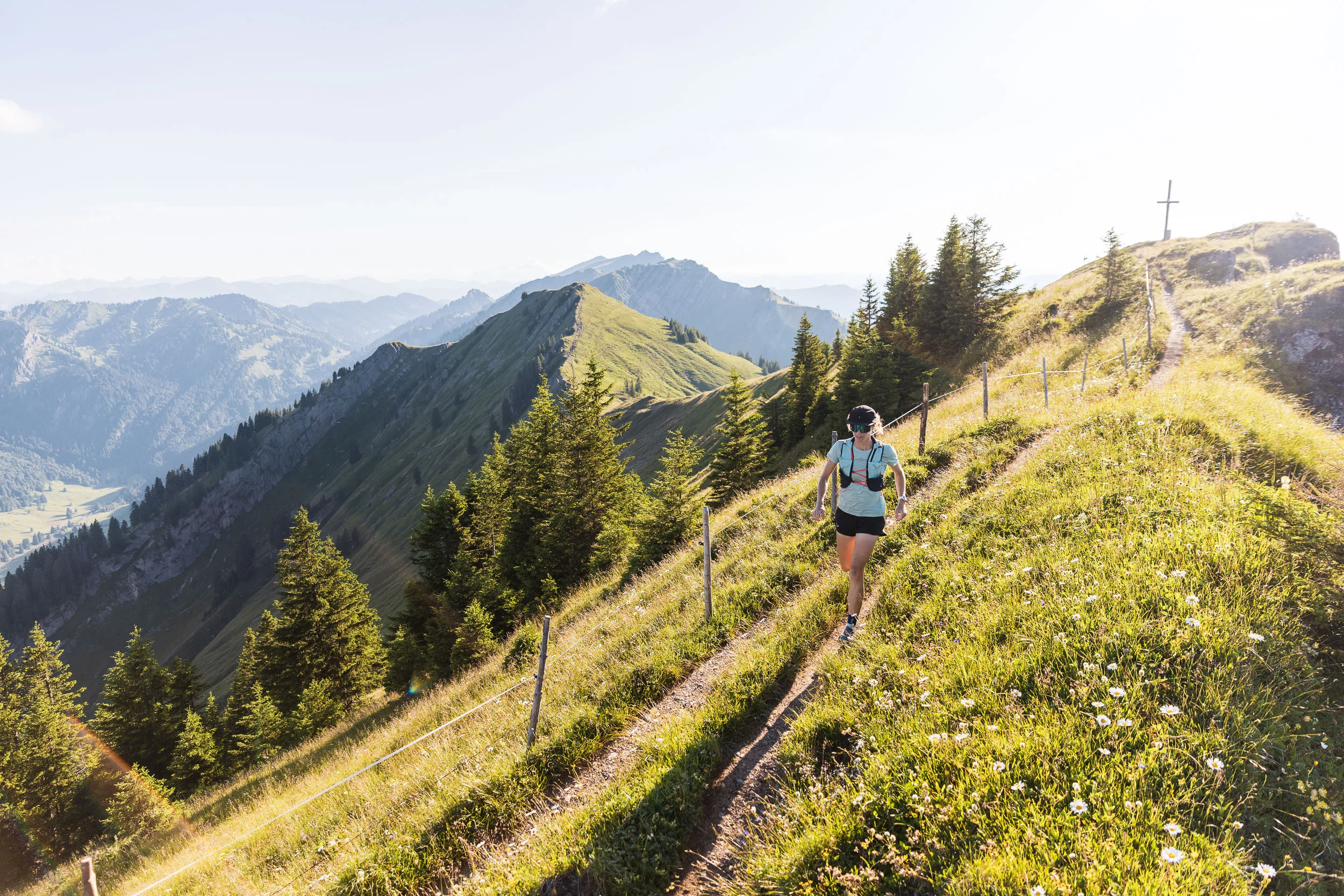 Eine Trailrunnerin läuft auf einem schmalen Pfad neben einer blühenden Bergwiese mit Blick auf grüne Grate.