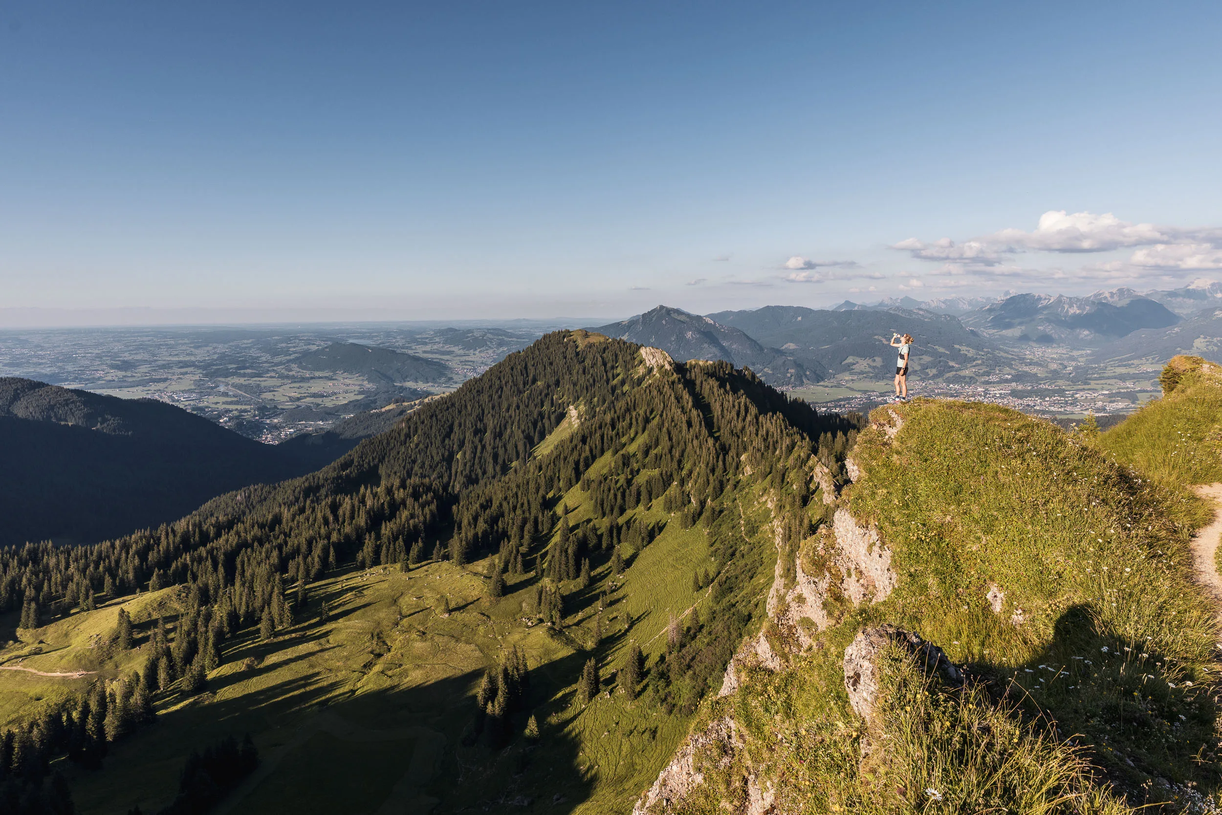 Eine Läuferin trinkt auf einer grasigen Felskante mit weitem Blick über Berge und das Allgäuer Tal.