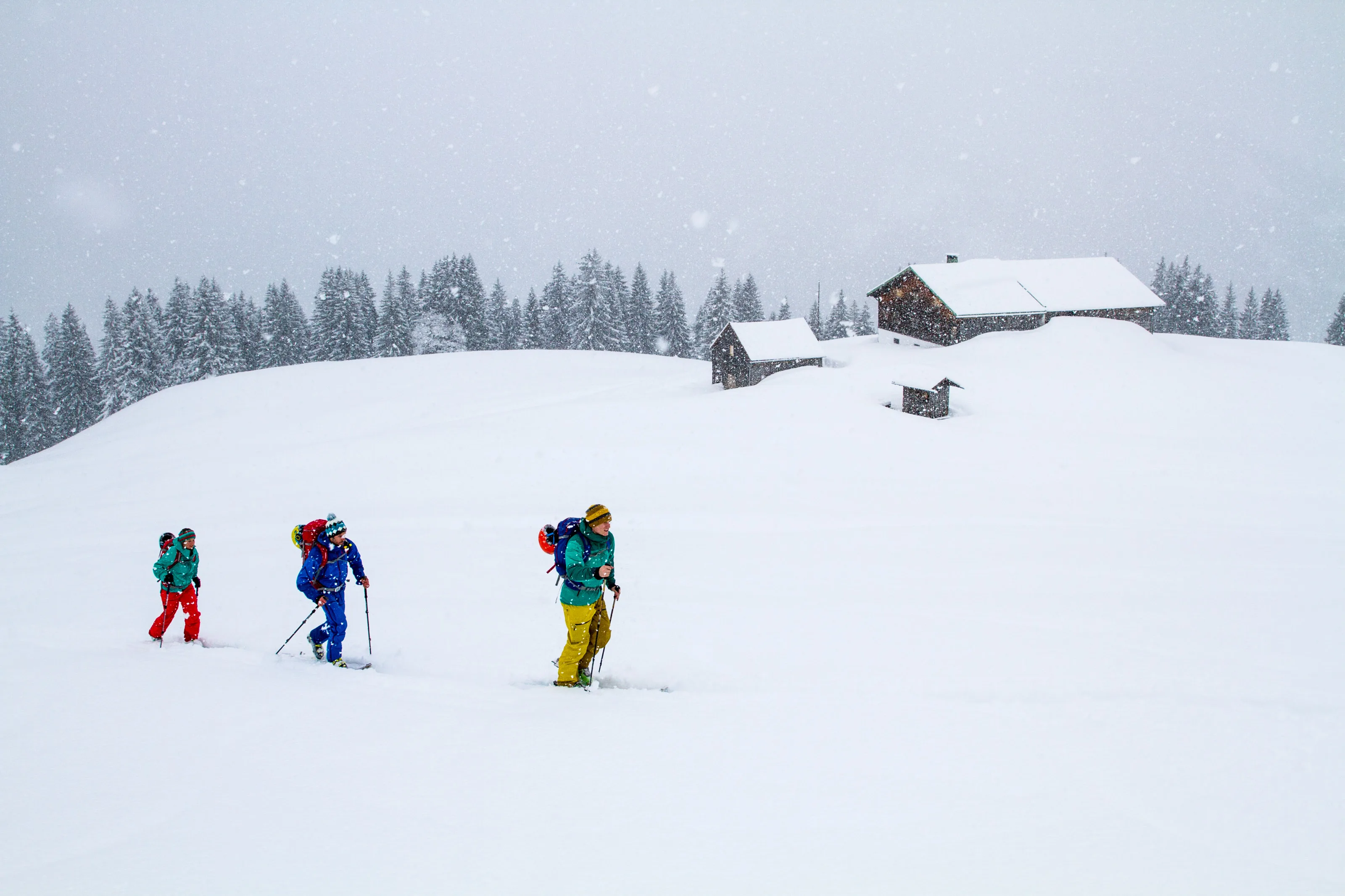 Vier Skitourengeher queren eine weite Schneefläche vor einer verschneiten Hütte