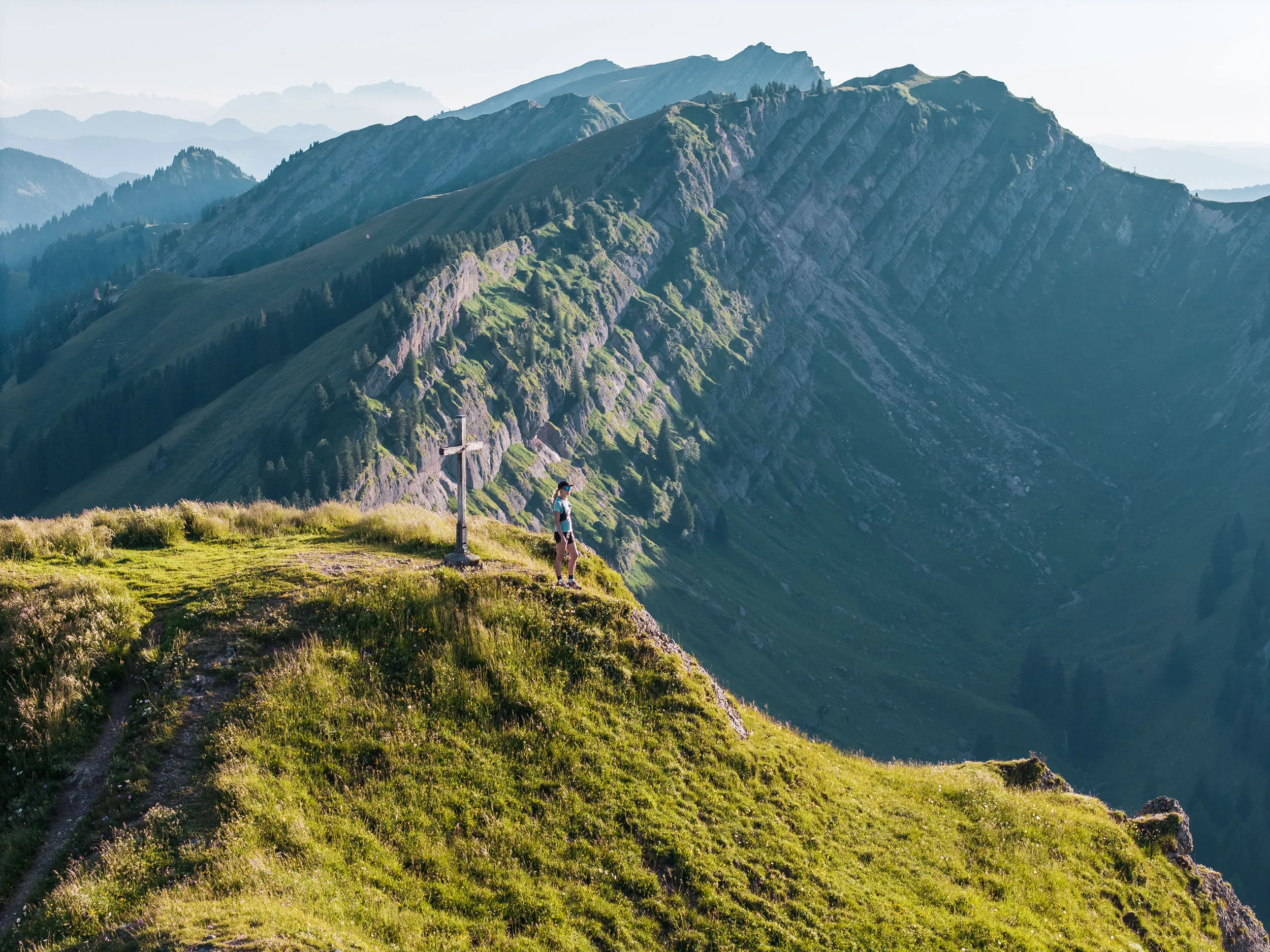 Eine Läuferin steht am Rand einer sonnigen Gipfelwiese vor einer steilen, schattigen Bergflanke.