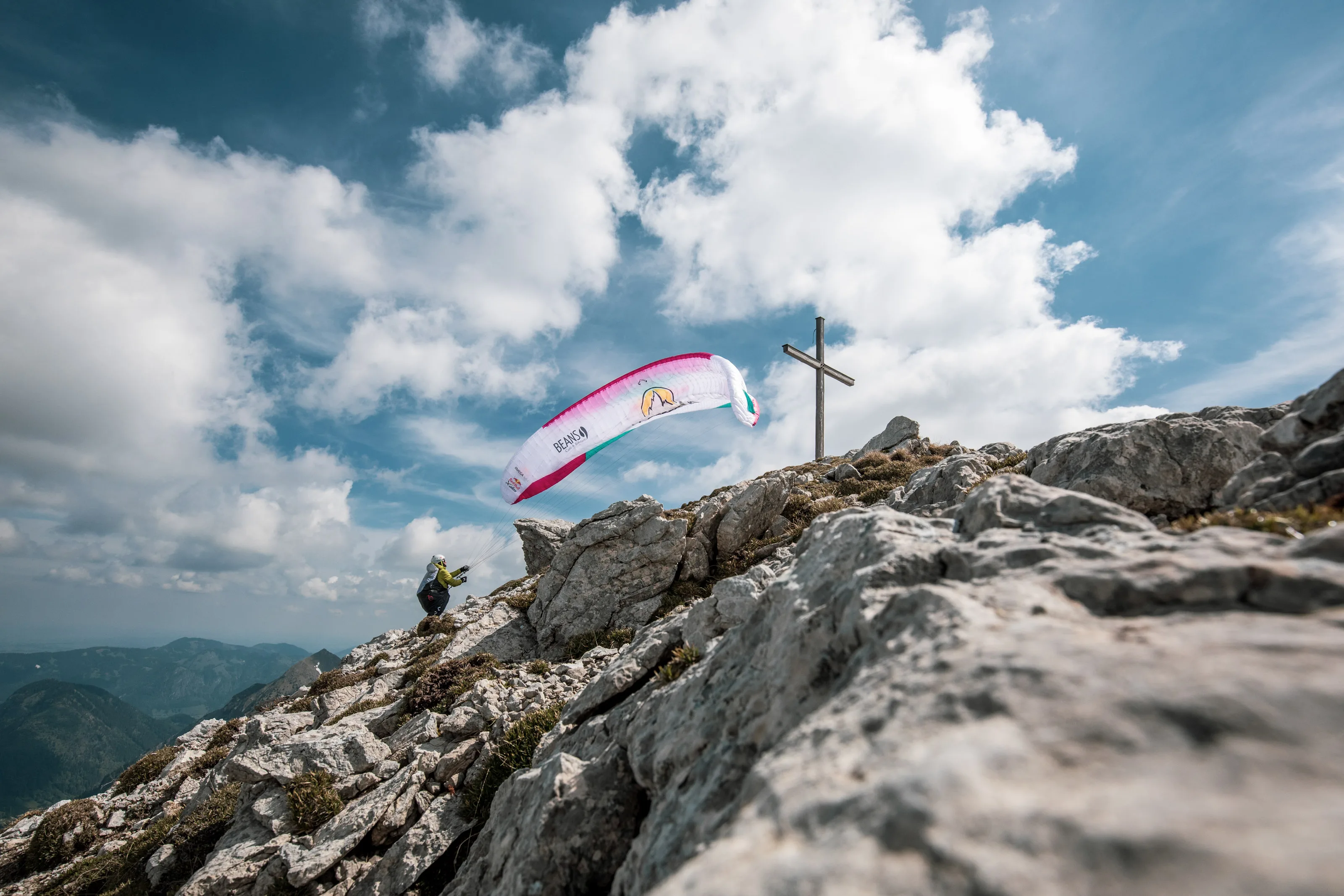 Ein pinkweißer Gleitschirm steigt hinter dem felsigen Grat und einem Gipfelkreuz in den Himmel.