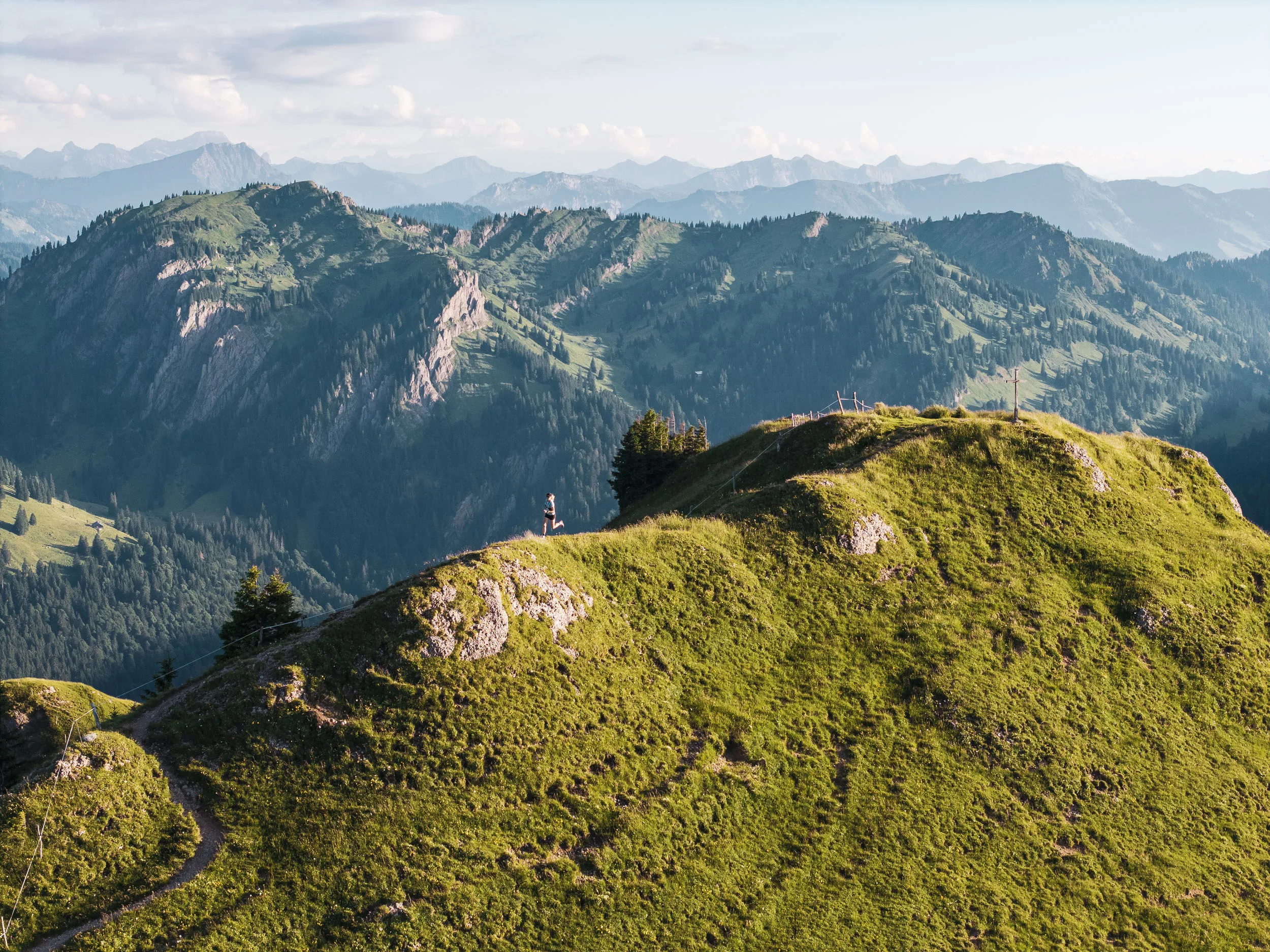 Eine Trailrunnerin läuft über einen grünen Grat, während das Gipfelkreuz hinter ihr zurückbleibt.