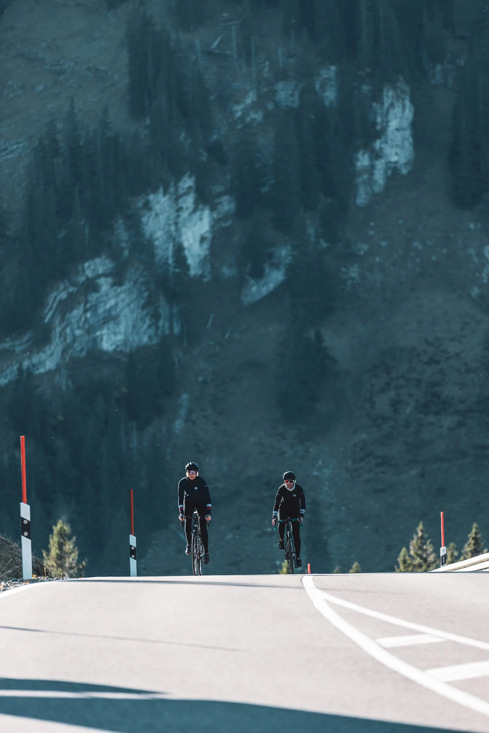 Zwei Rennradfahrerinnen fahren vor einer dunklen Felswand bergauf