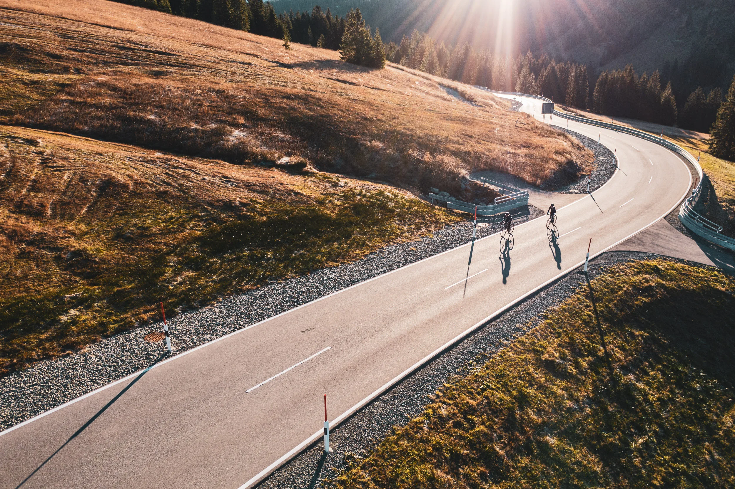Zwei Rennradfahrerinnen auf einer geschwungenen Passstraße im warmen Herbstlicht