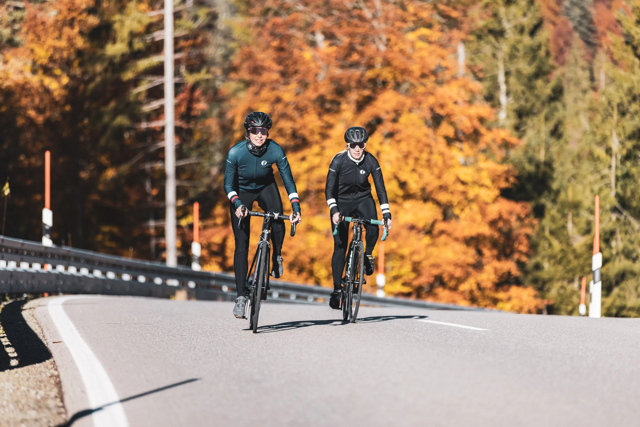 Zwei Rennradfahrerinnen auf der Passstraße vor leuchtendem Herbstwald