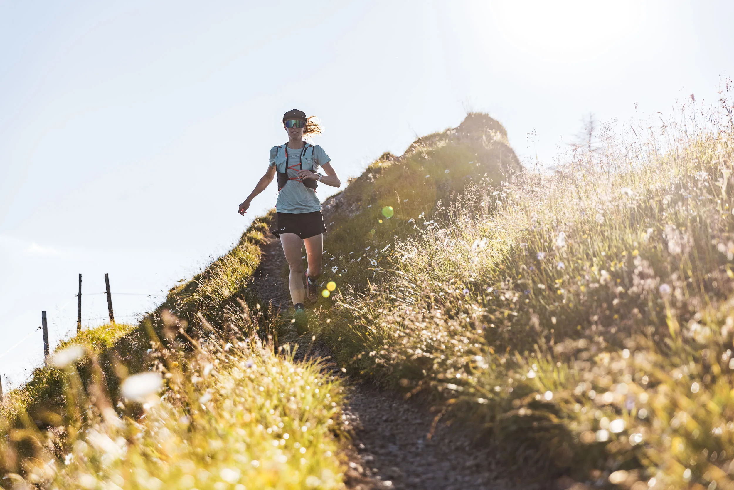 Eine Trailrunnerin läuft einen schmalen Pfad zwischen hell beleuchteten Gräsern und Bergblumen hinab.