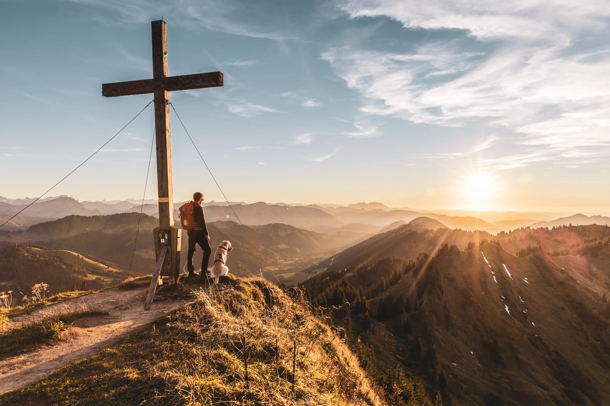 Wanderer und Hund am Gipfelkreuz vor der tief stehenden Sonne im Oberallgäu