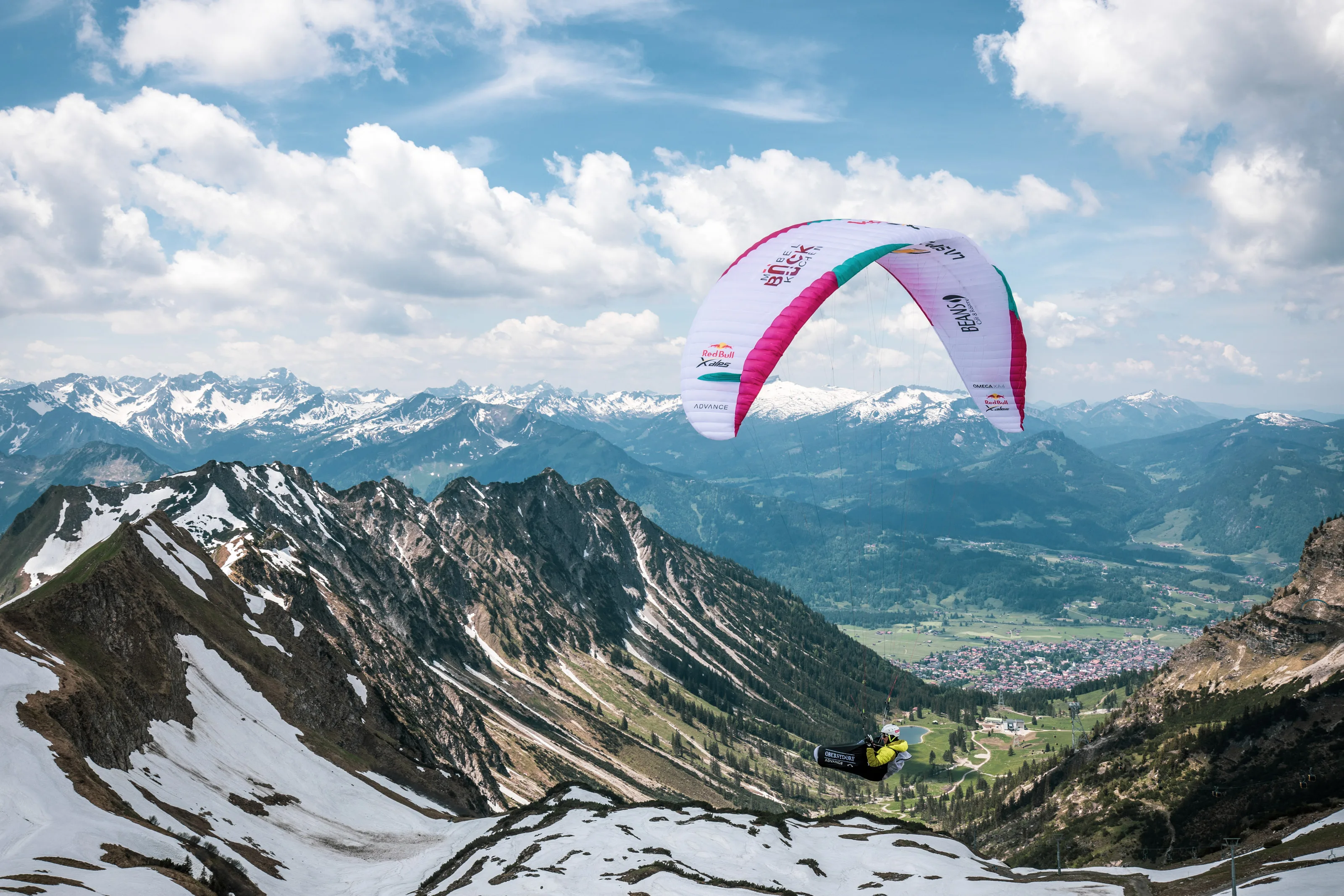 Michael Lacher fliegt unter dem pinkweißen Gleitschirm über einem Tal zwischen dunklen Bergen.