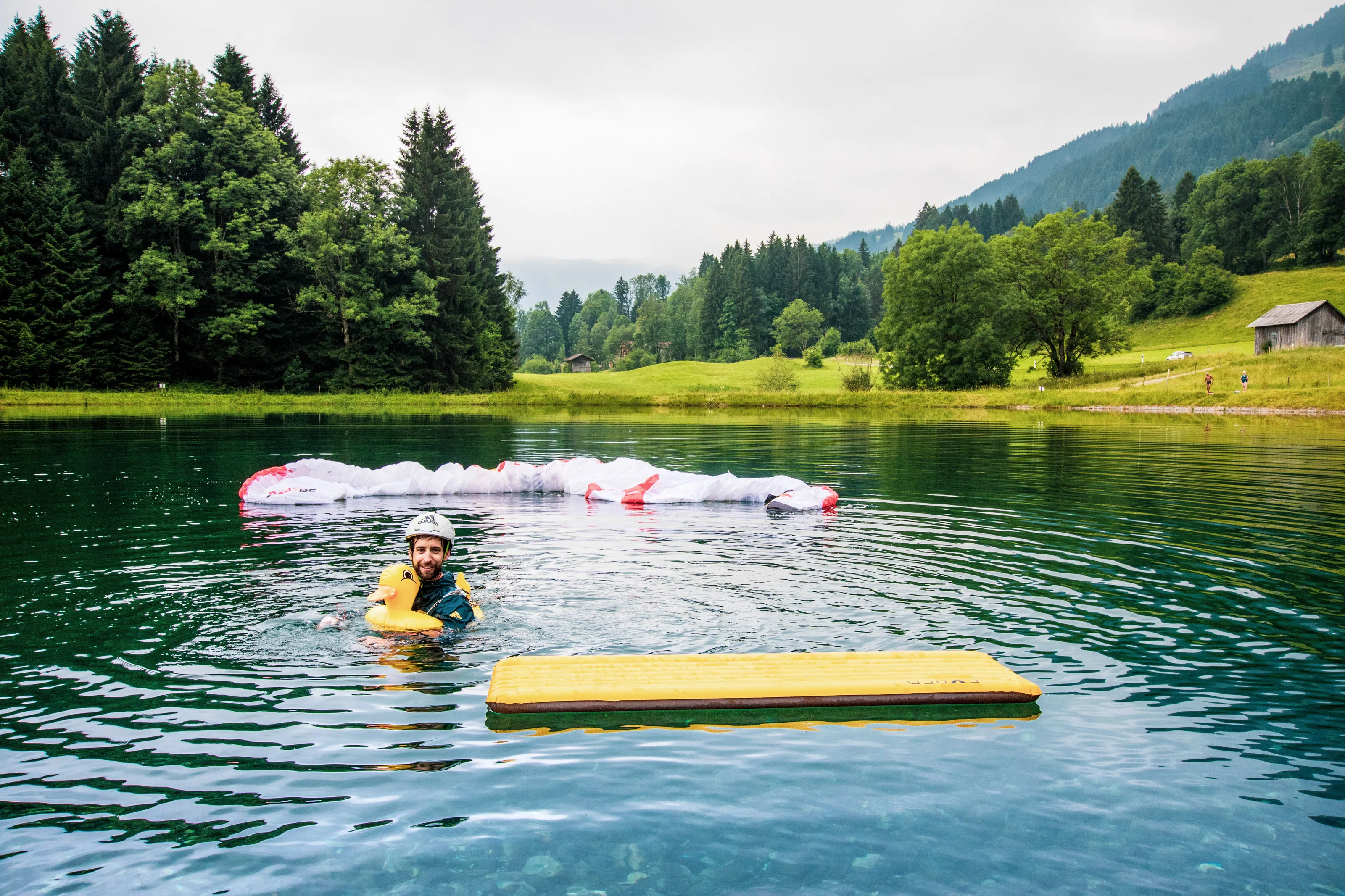 Manuel Nübel schwimmt mit einer gelben Badeente im Teich; sein Gleitschirm treibt hinter ihm auf dem Wasser.