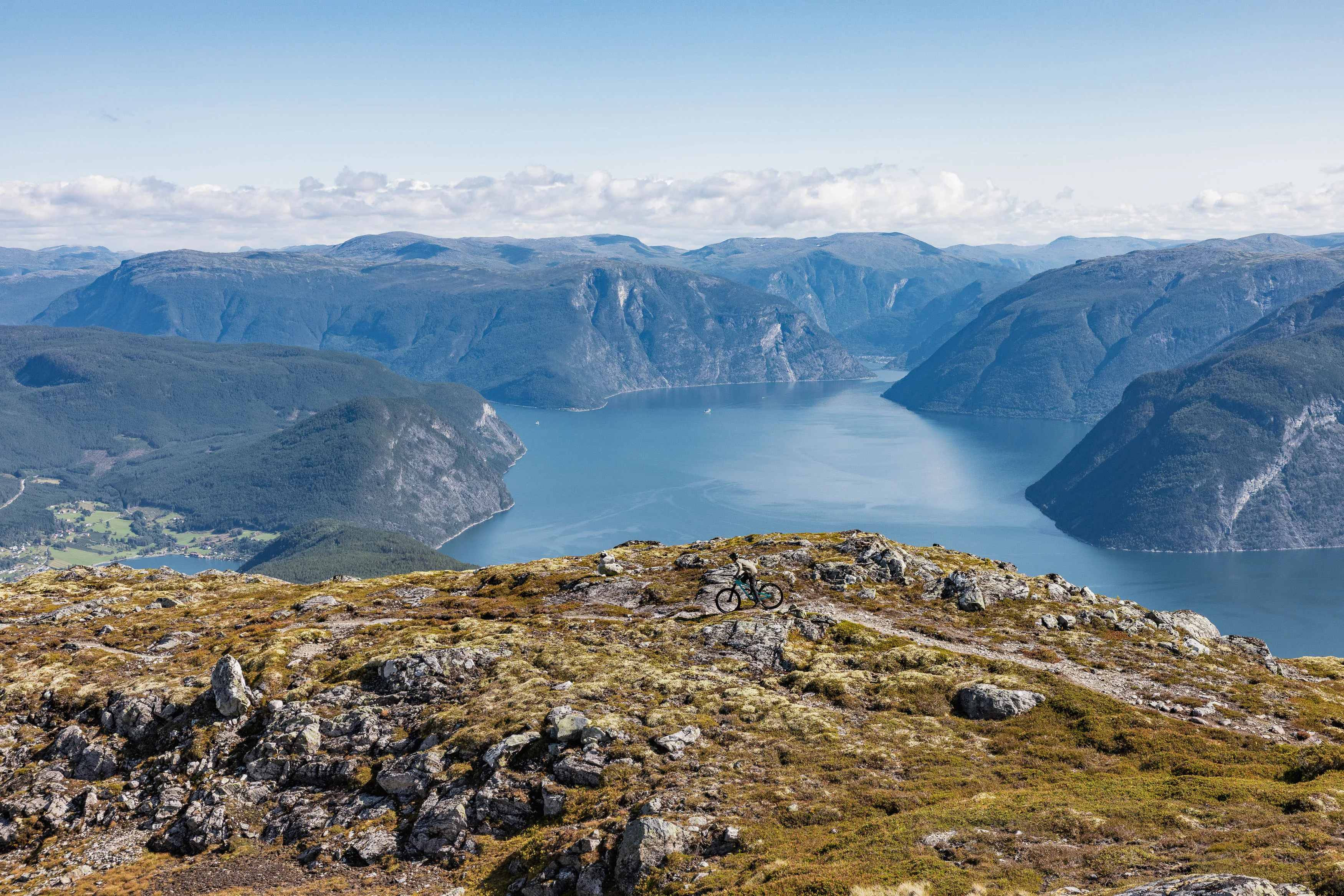Mountainbiker fährt klein im Bild über eine offene Hochebene mit weitem Fjordpanorama