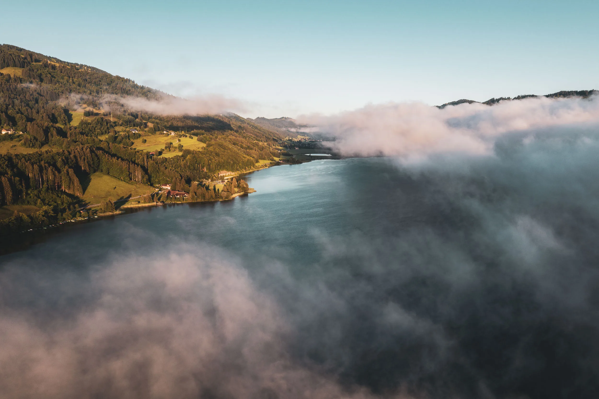 Luftaufnahme des Alpsees mit herbstlichen Ufern und Wolken über der Wasseroberfläche.