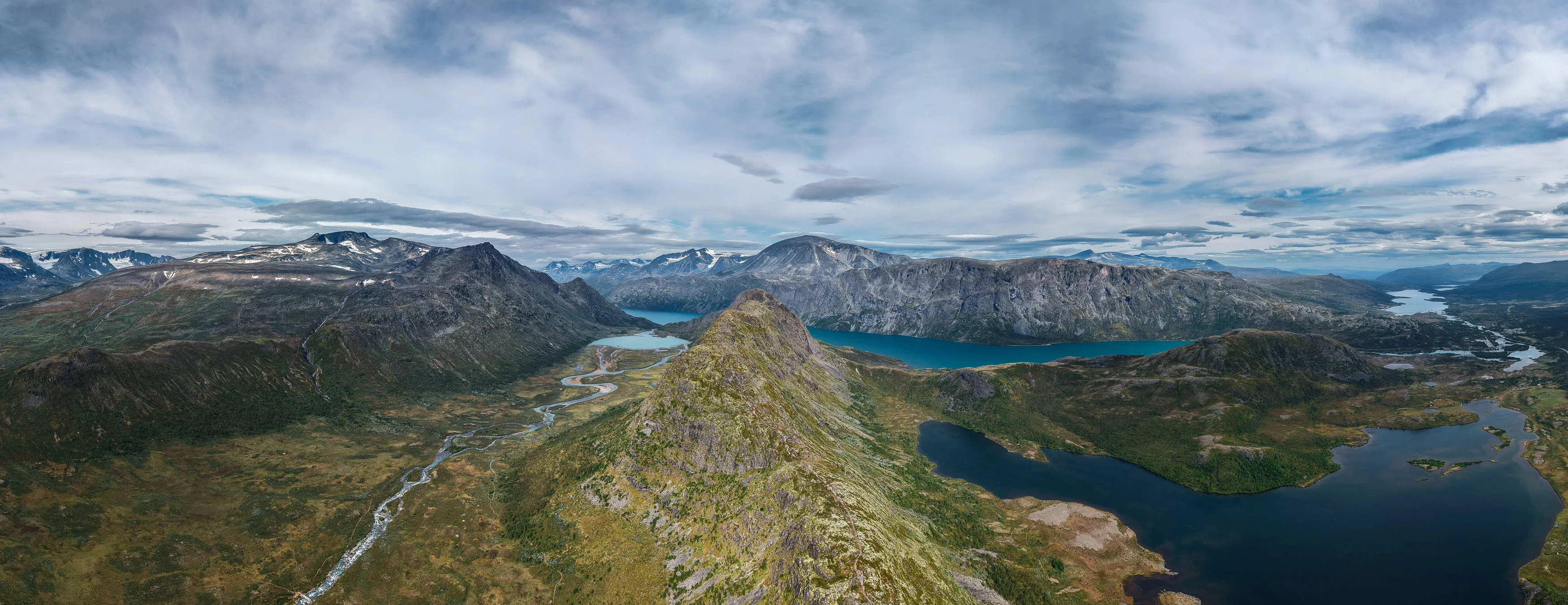 Weites Luftpanorama mit Seen, Bergrücken und Schneefeldern unter bewegtem Himmel