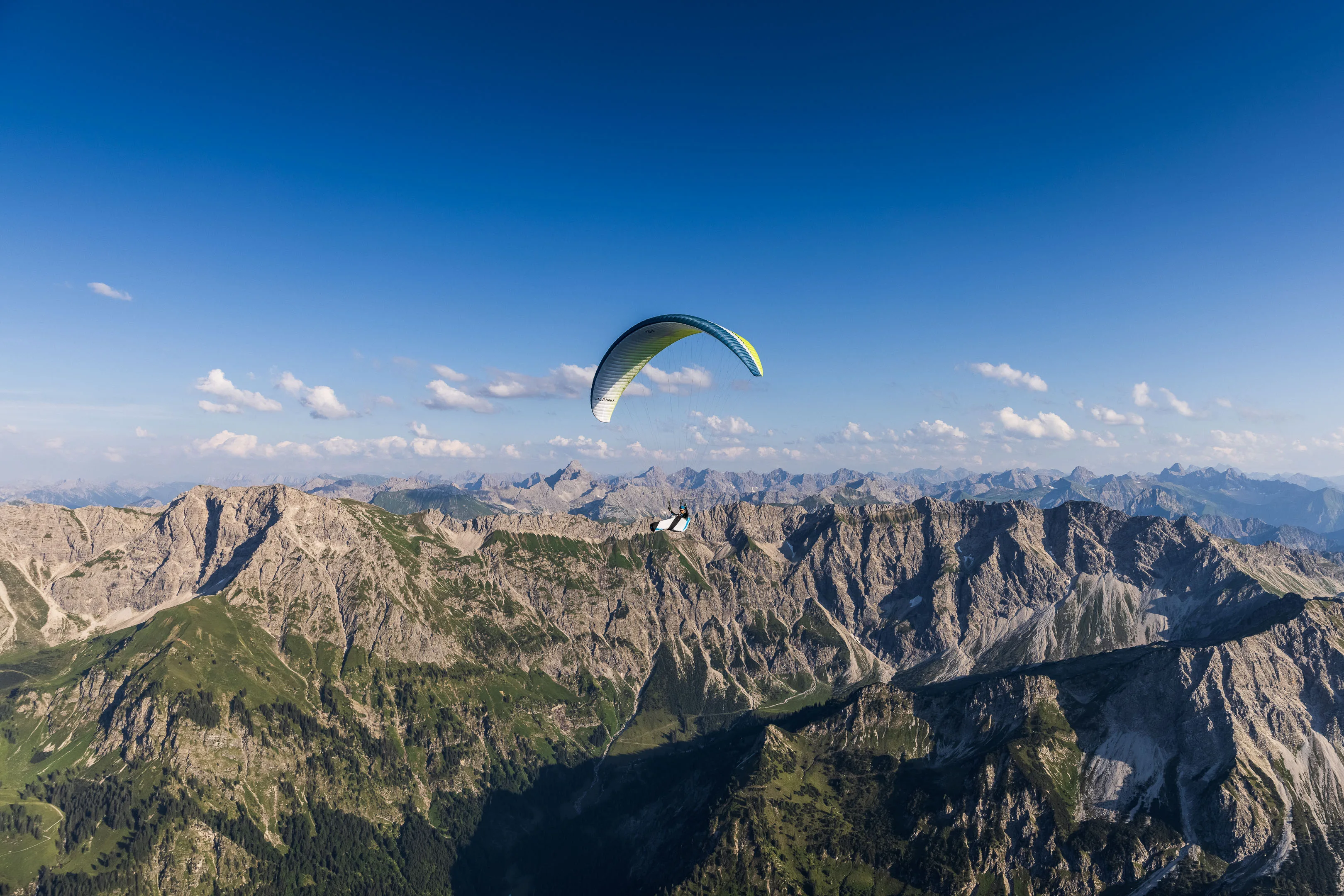 Ein Gleitschirmpilot fliegt hoch über einem dunklen Tal vor fernen Bergketten