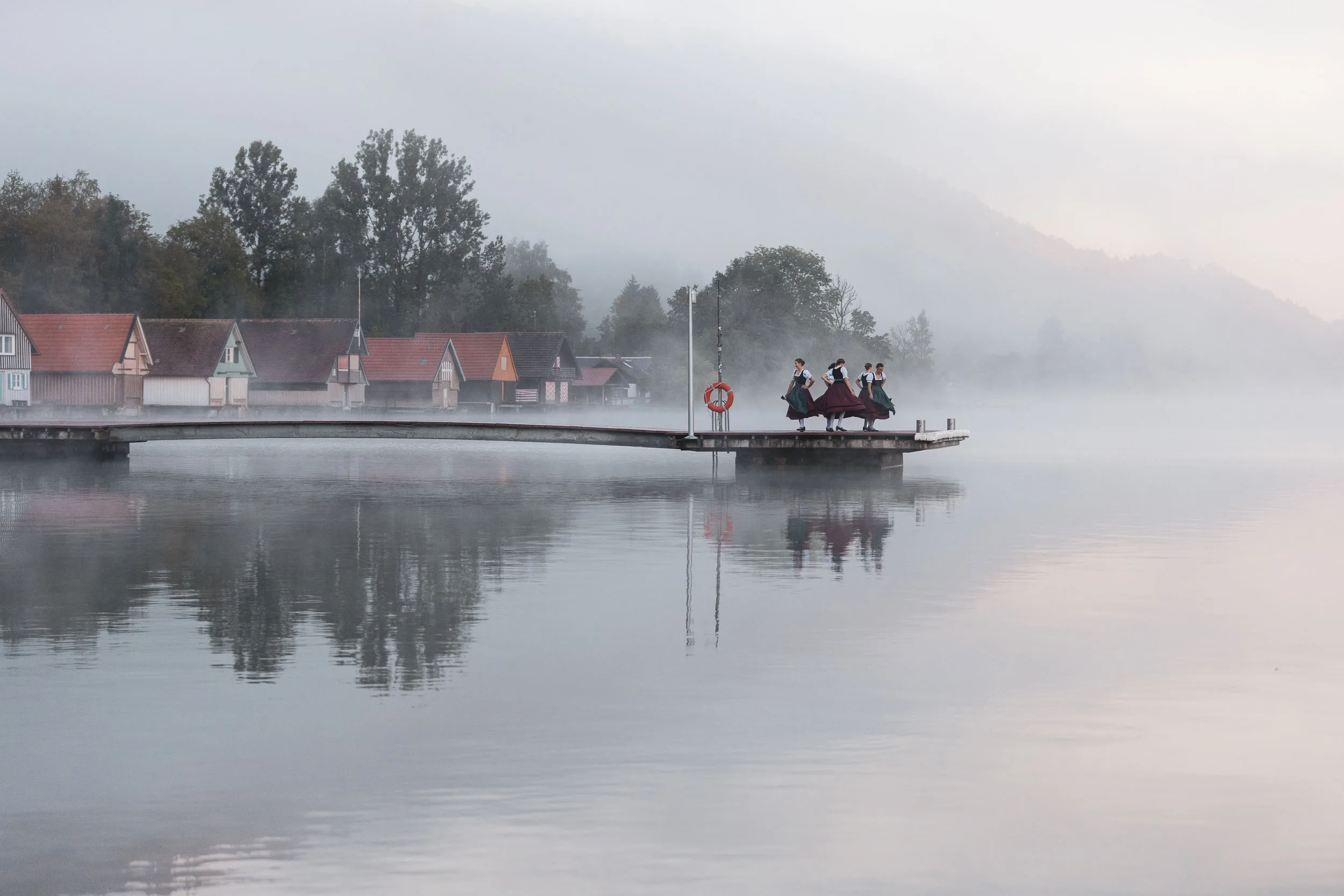 Tänzerinnen in Tracht auf einem Steg am stillen Alpsee im Morgennebel.