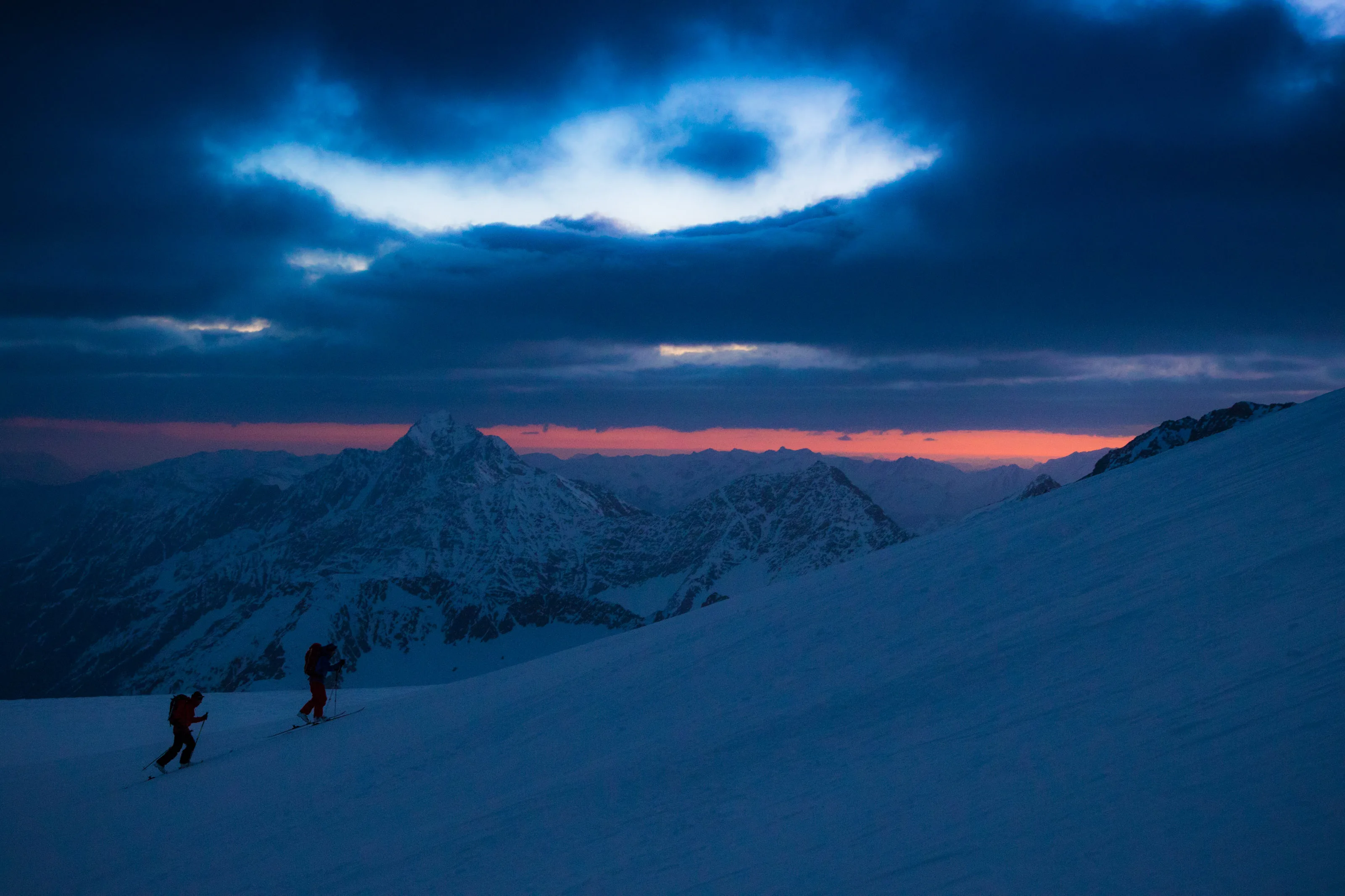 Zwei Skitourengeher folgen einer Spur über einen breiten Schneehang unter dunklen Wolken