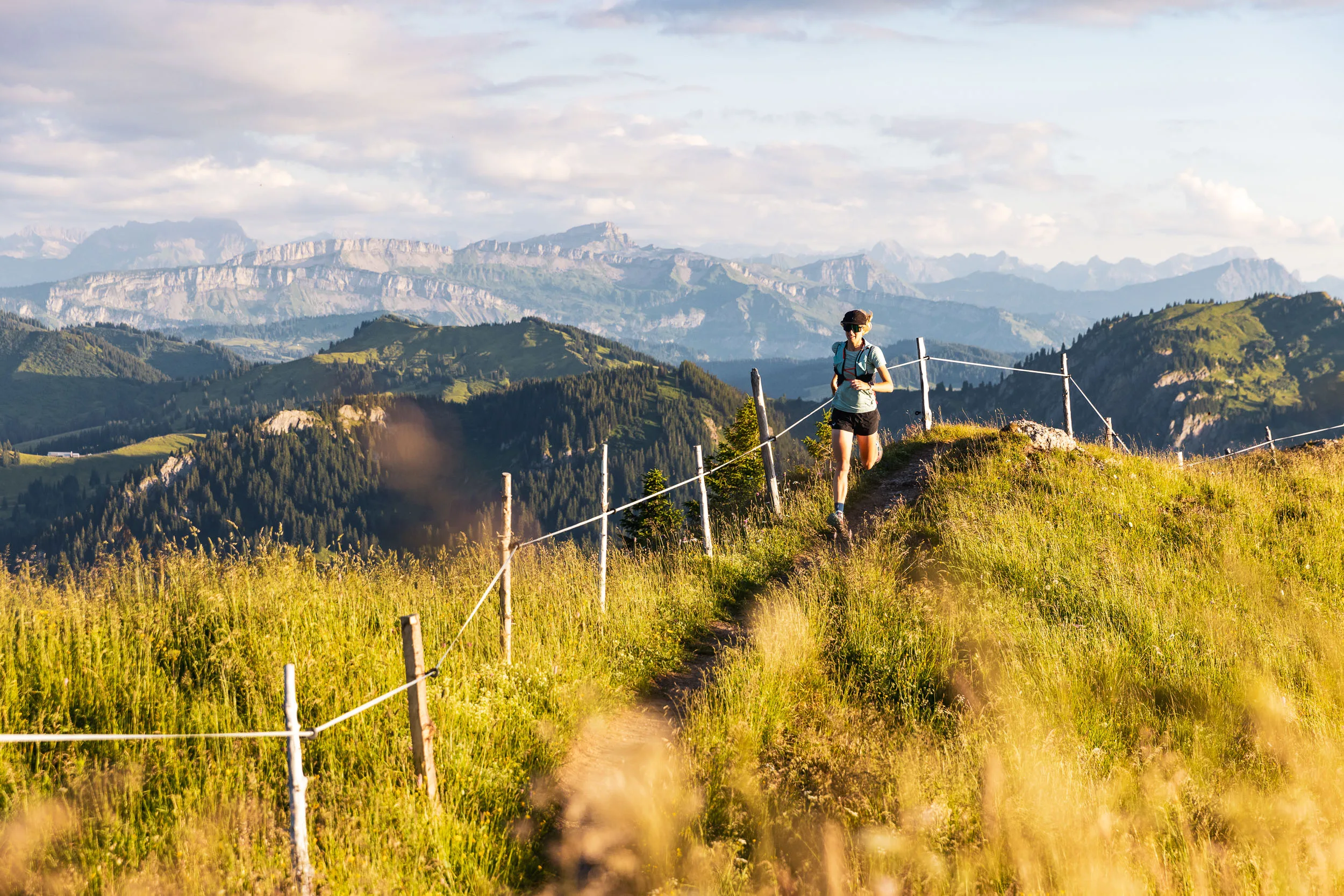 Eine Trailrunnerin folgt einem schmalen Höhenweg zwischen goldenen Gräsern vor gestaffelten Bergketten.