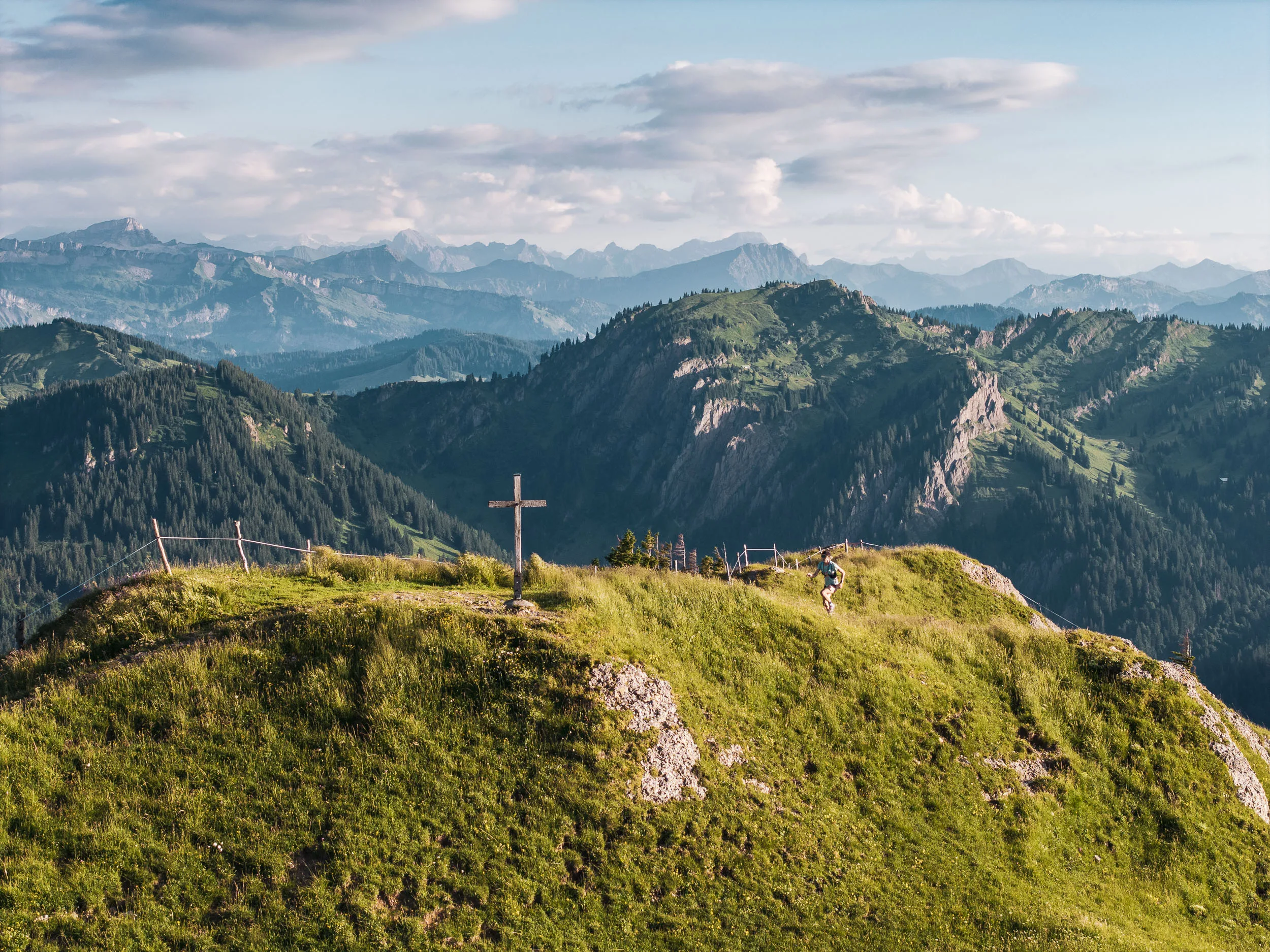 Eine Trailrunnerin erreicht einen grasbewachsenen Gipfel mit Holzkreuz vor einem weiten Bergpanorama.