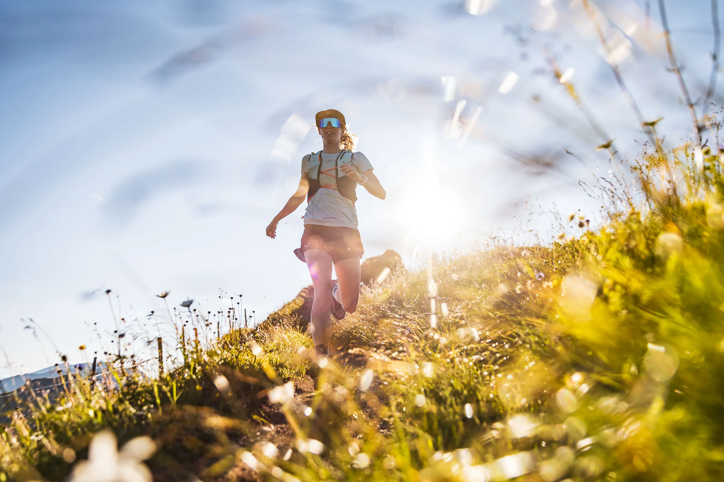 Eine Trailrunnerin läuft im Gegenlicht bergab, unscharfe Gräser und Blüten rahmen den Vordergrund.