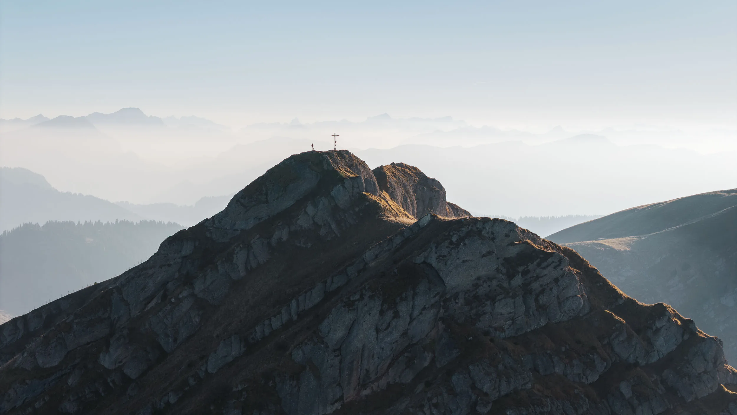 Der felsige Gipfel mit Kreuz hebt sich vor einer Kette ferner Alpen ab