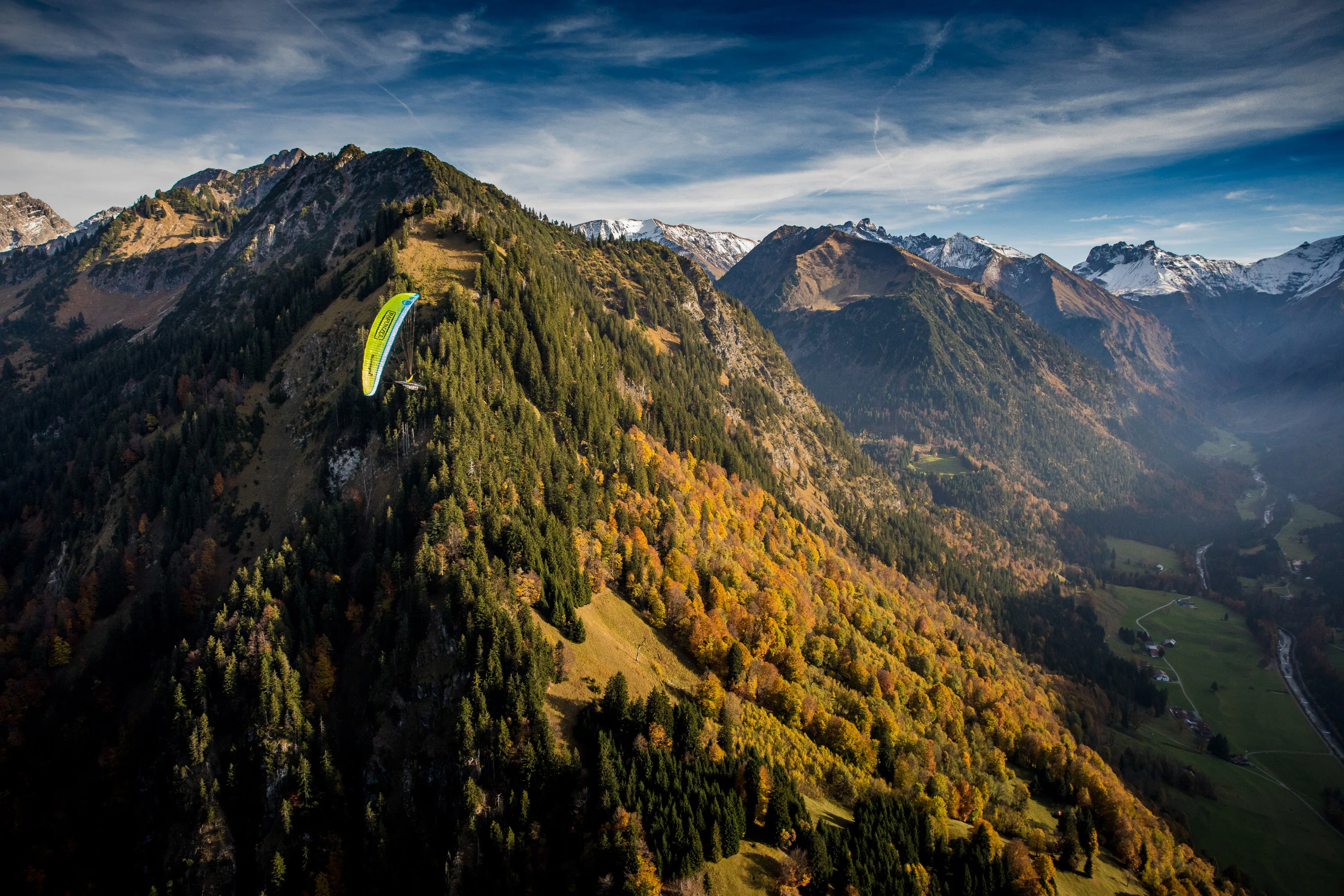 Michael Gebert fliegt mit grünem Gleitschirm entlang eines bewaldeten Herbsthangs über dem Oberstdorfer Tal