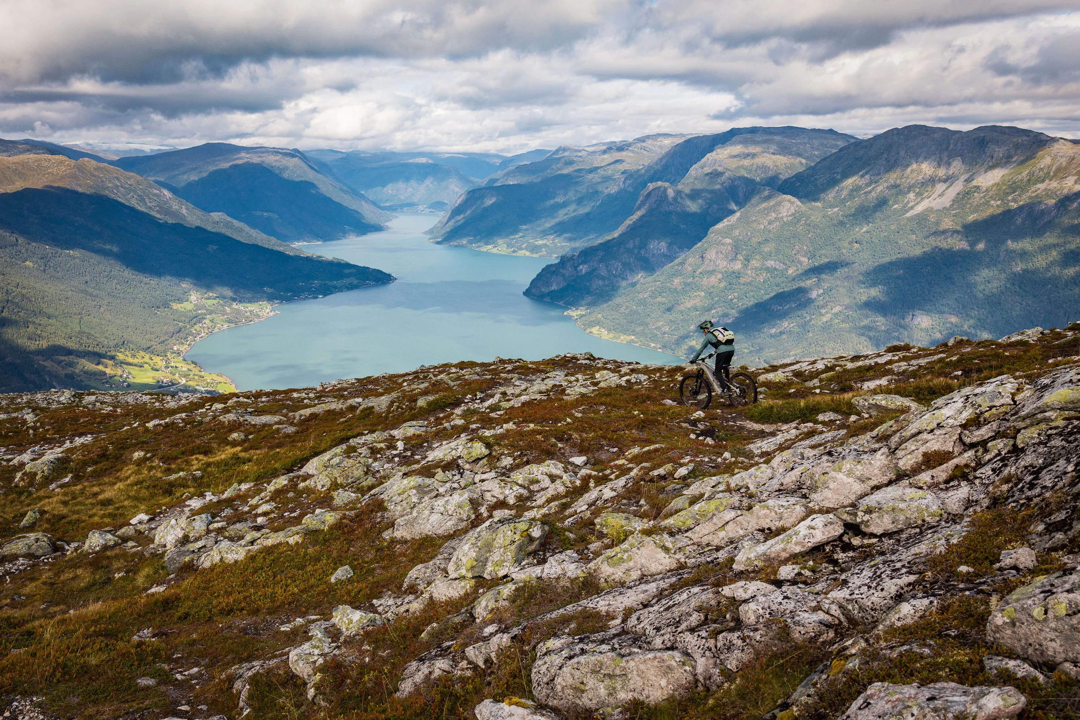 Mountainbiker quert einen felsigen Hang mit Fjorden und Bergen im Hintergrund