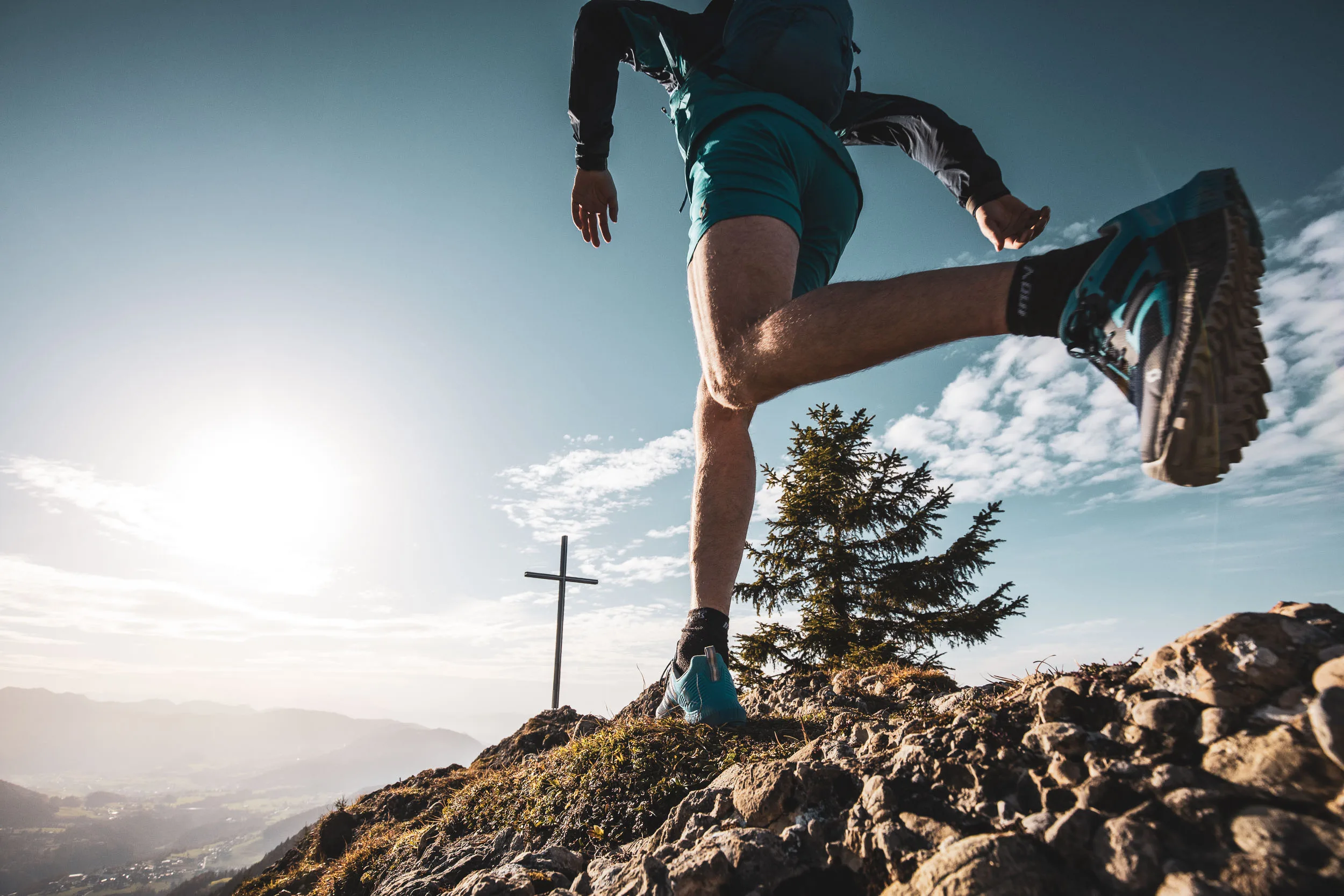 Andreas Kögel setzt beim Trailrunning vor einem Gipfelkreuz zum Sprung über felsigen Boden an.