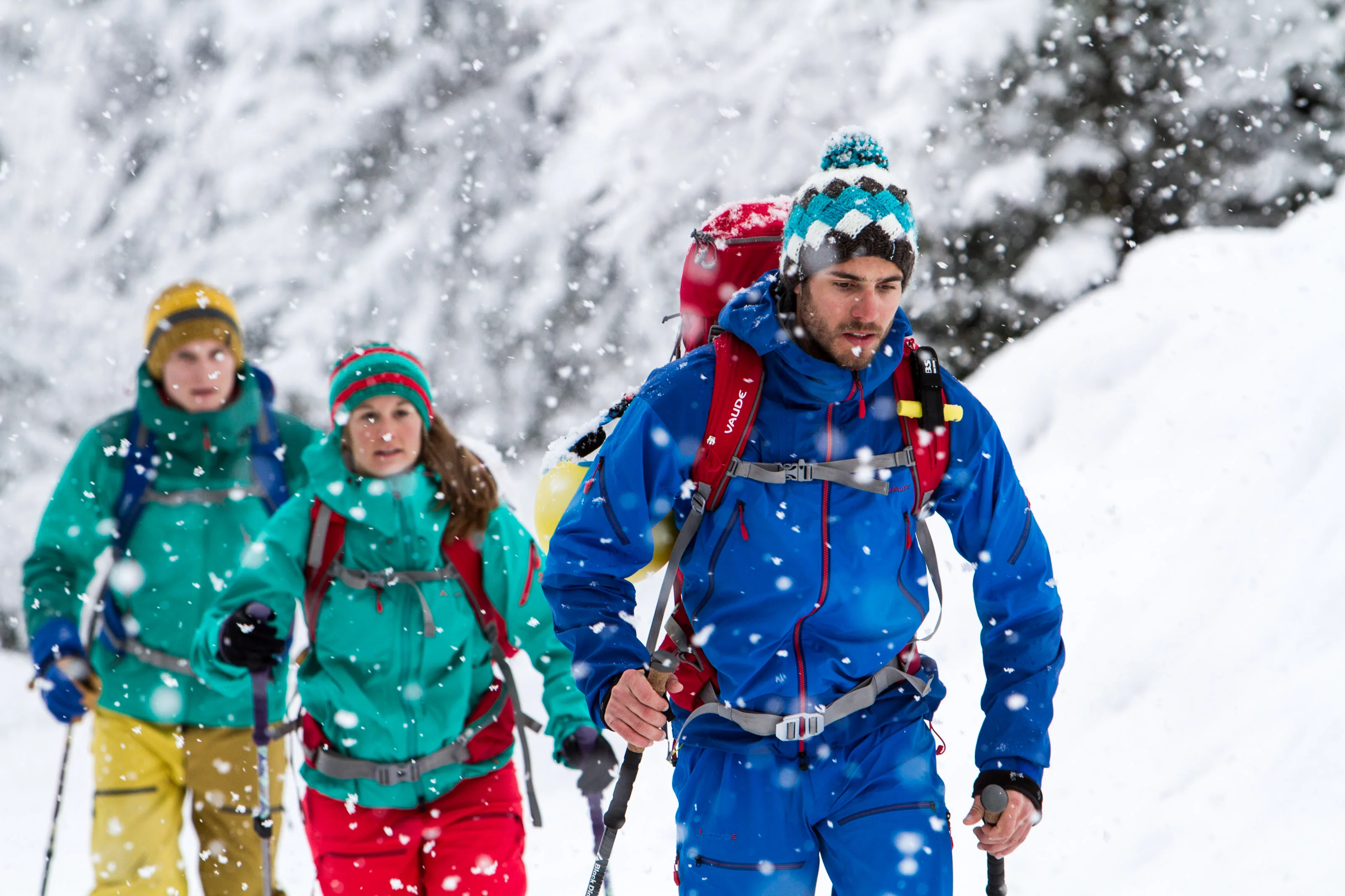 Drei Skitourengeher mit Rucksäcken im dichten Schneefall vor verschneiten Bäumen