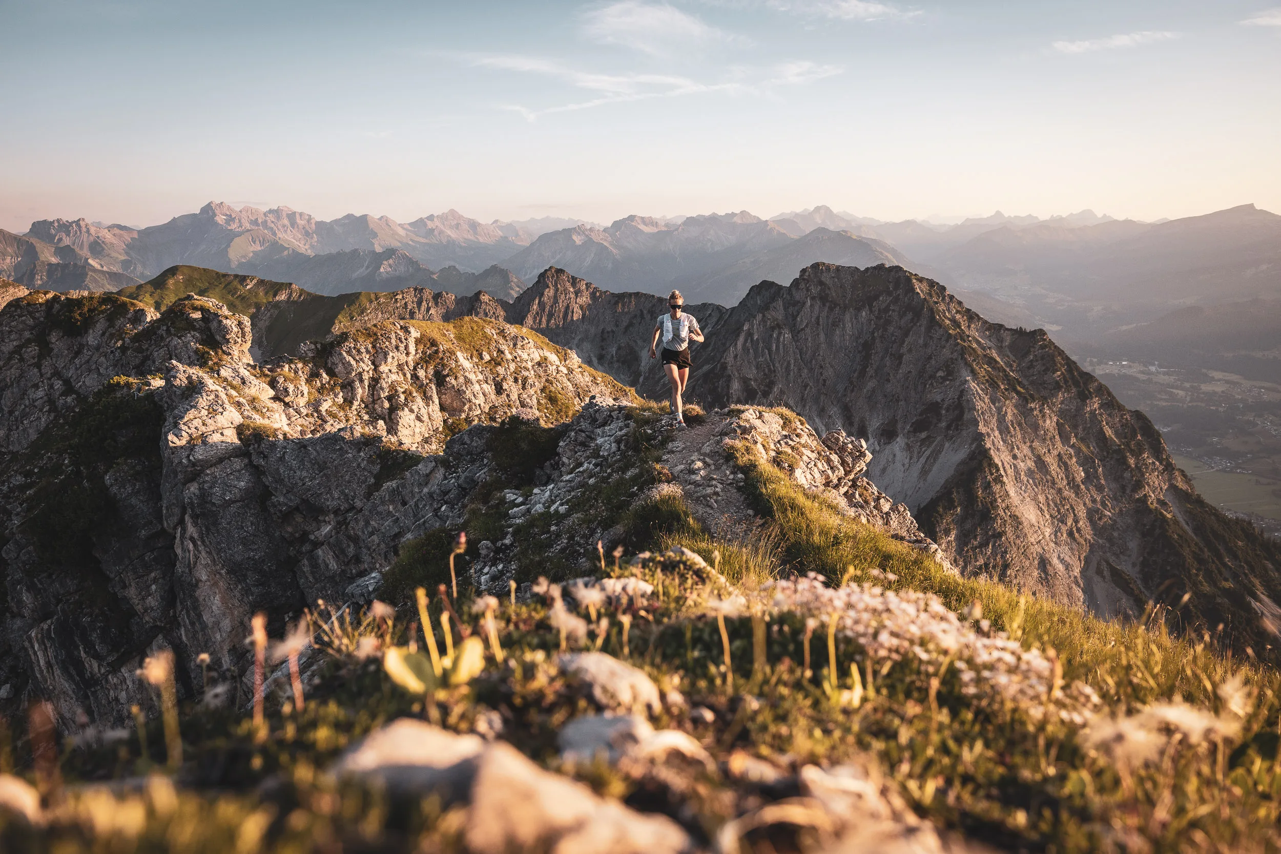 Trailrunnerin zwischen Bergblumen auf einem felsigen Grat im Abendlicht