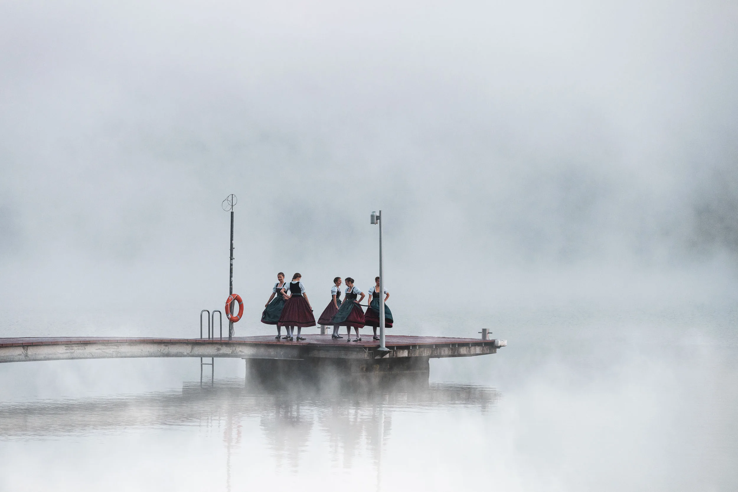 Tänzerinnen in Tracht auf einem Steg am Alpsee vor dichter Nebelkulisse.