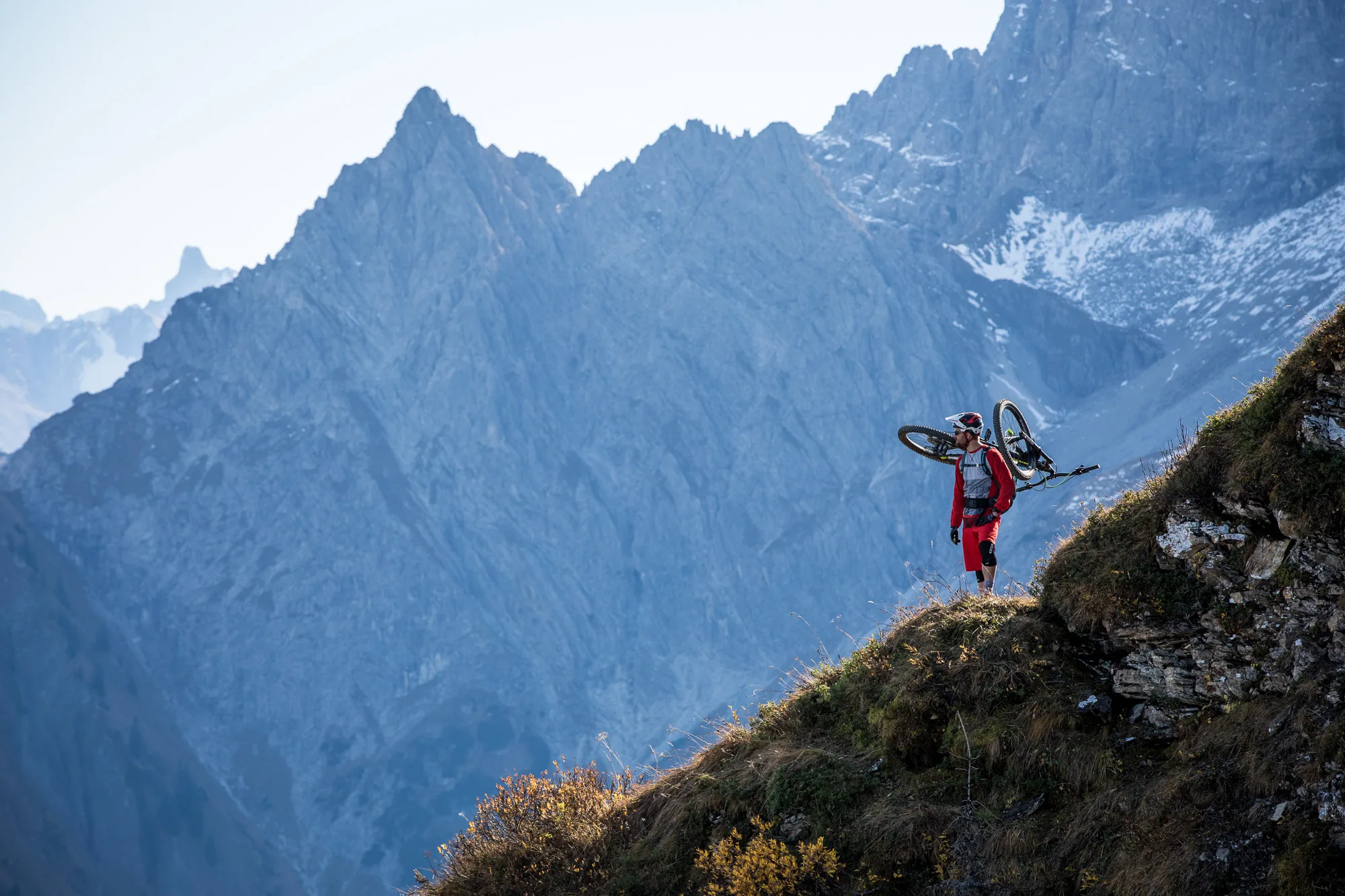 Mountainbiker trägt sein Rad über einen Grat vor einer schroffen alpinen Kulisse