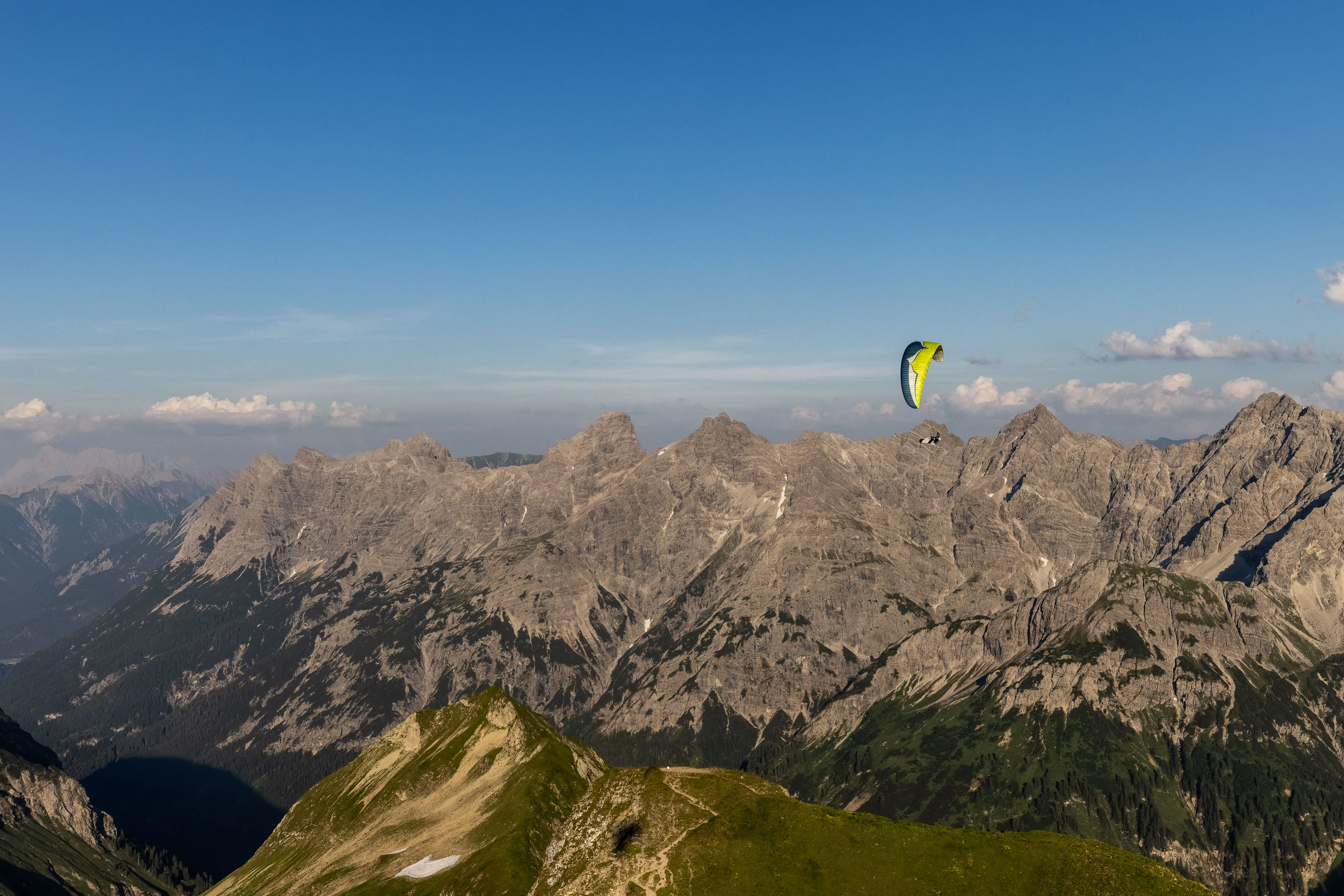 Ein gelber Gleitschirm fliegt vor einer breiten grauen Felswand über grüne Hänge
