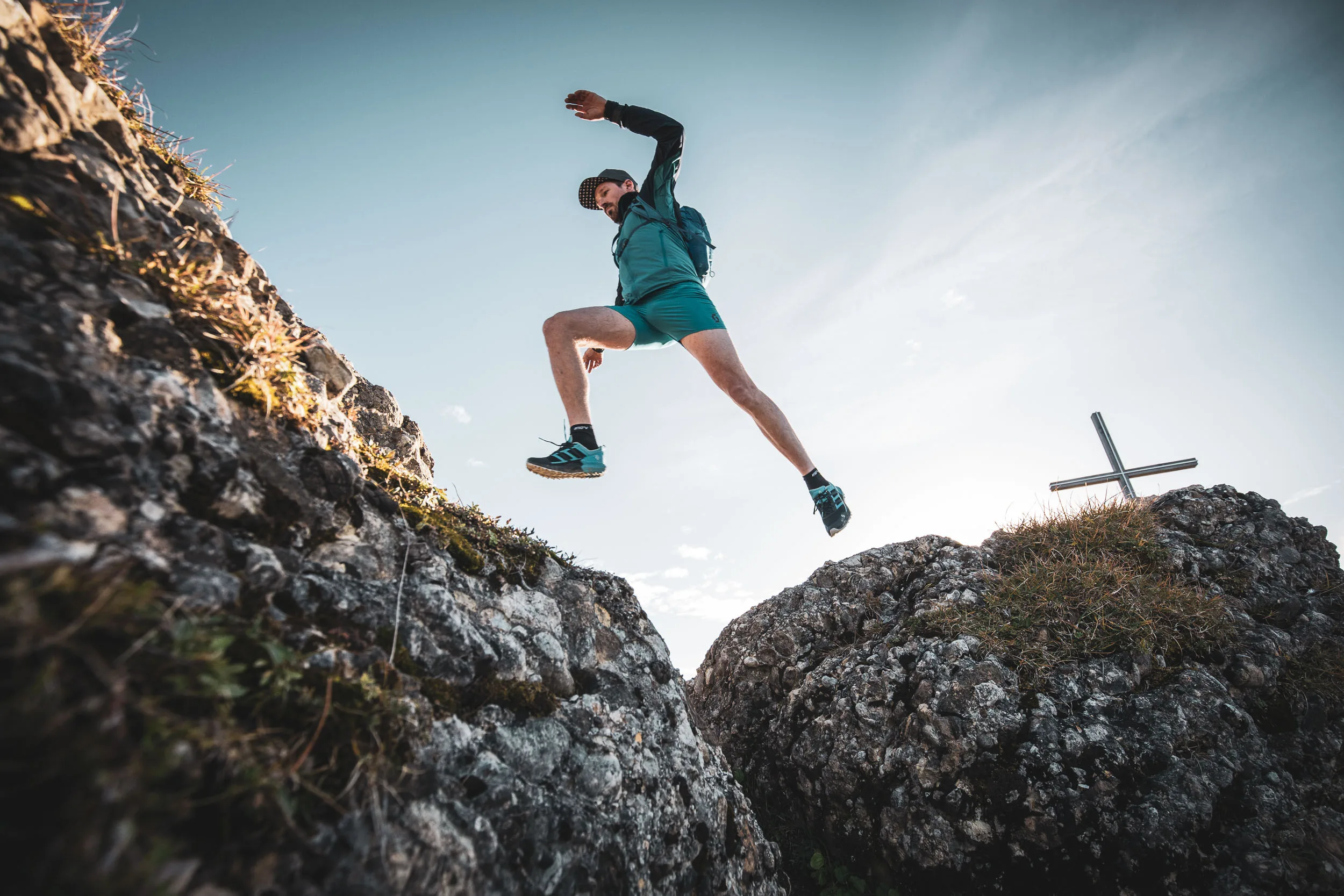 Andreas Kögel springt beim Trailrunning zwischen zwei Felsen an einem Gipfelkreuz.