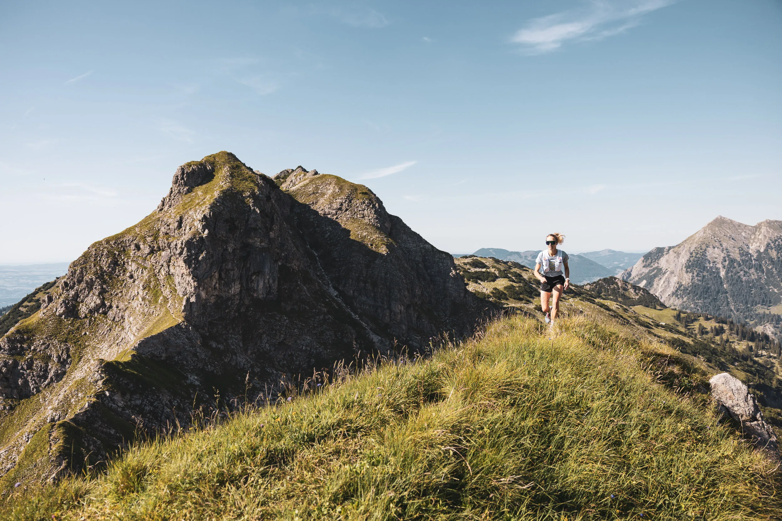 Trailrunnerin auf einem grasigen Berggrat vor einem markanten Felsgipfel