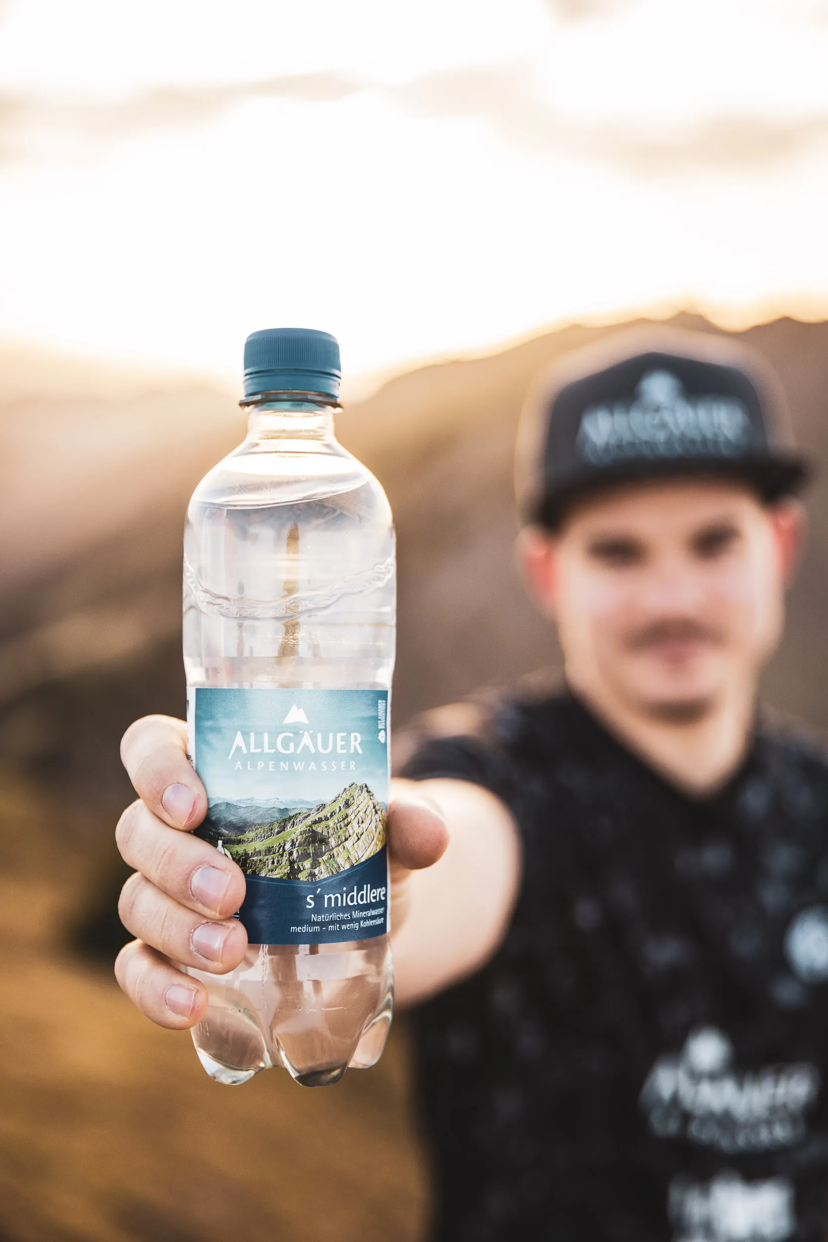 Ausgestreckte Hand haelt eine Mineralwasserflasche vor einem unscharfen Trailrunner