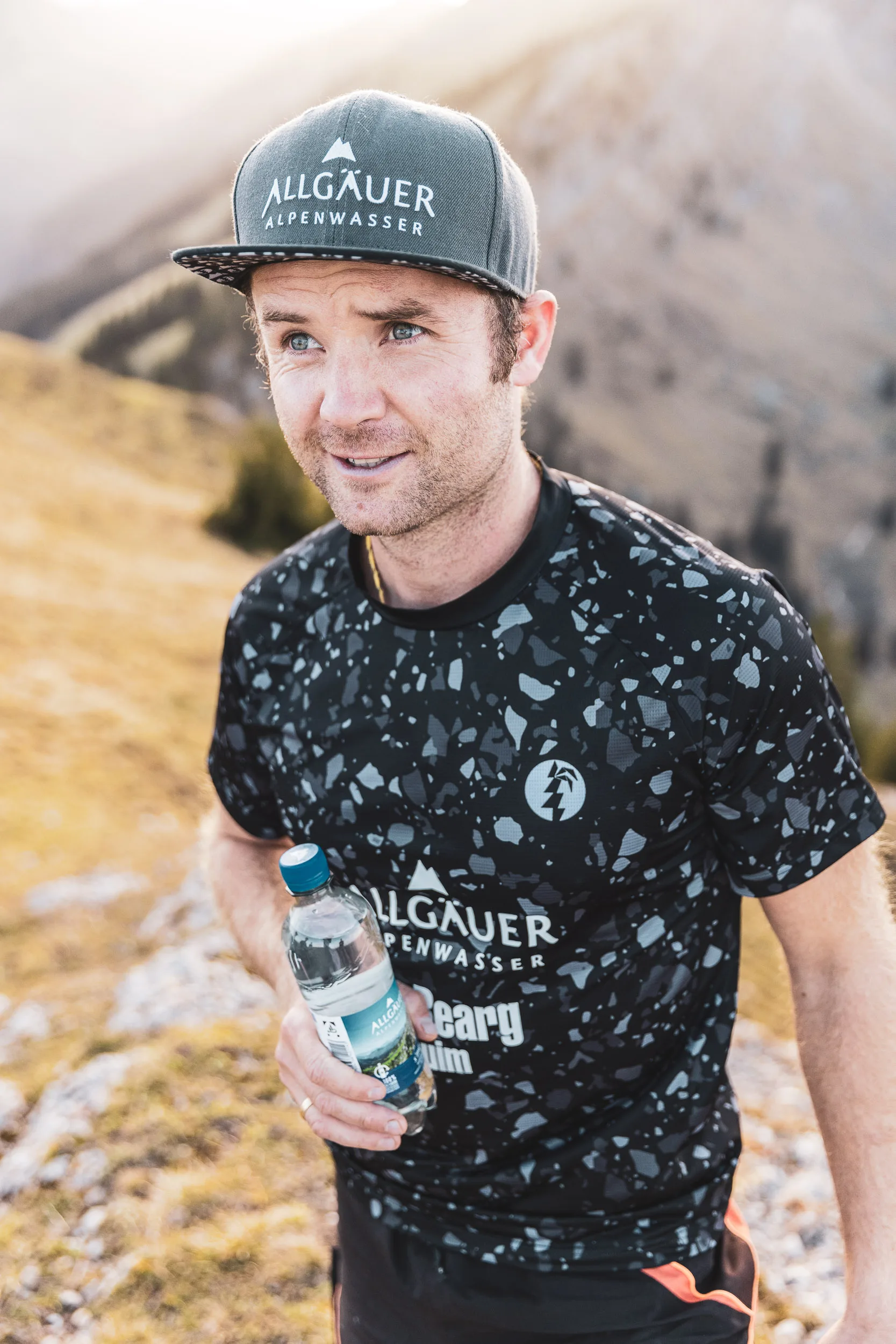 Trailrunner steht mit Wasserflasche auf alpinem Pfad vor Bergkulisse