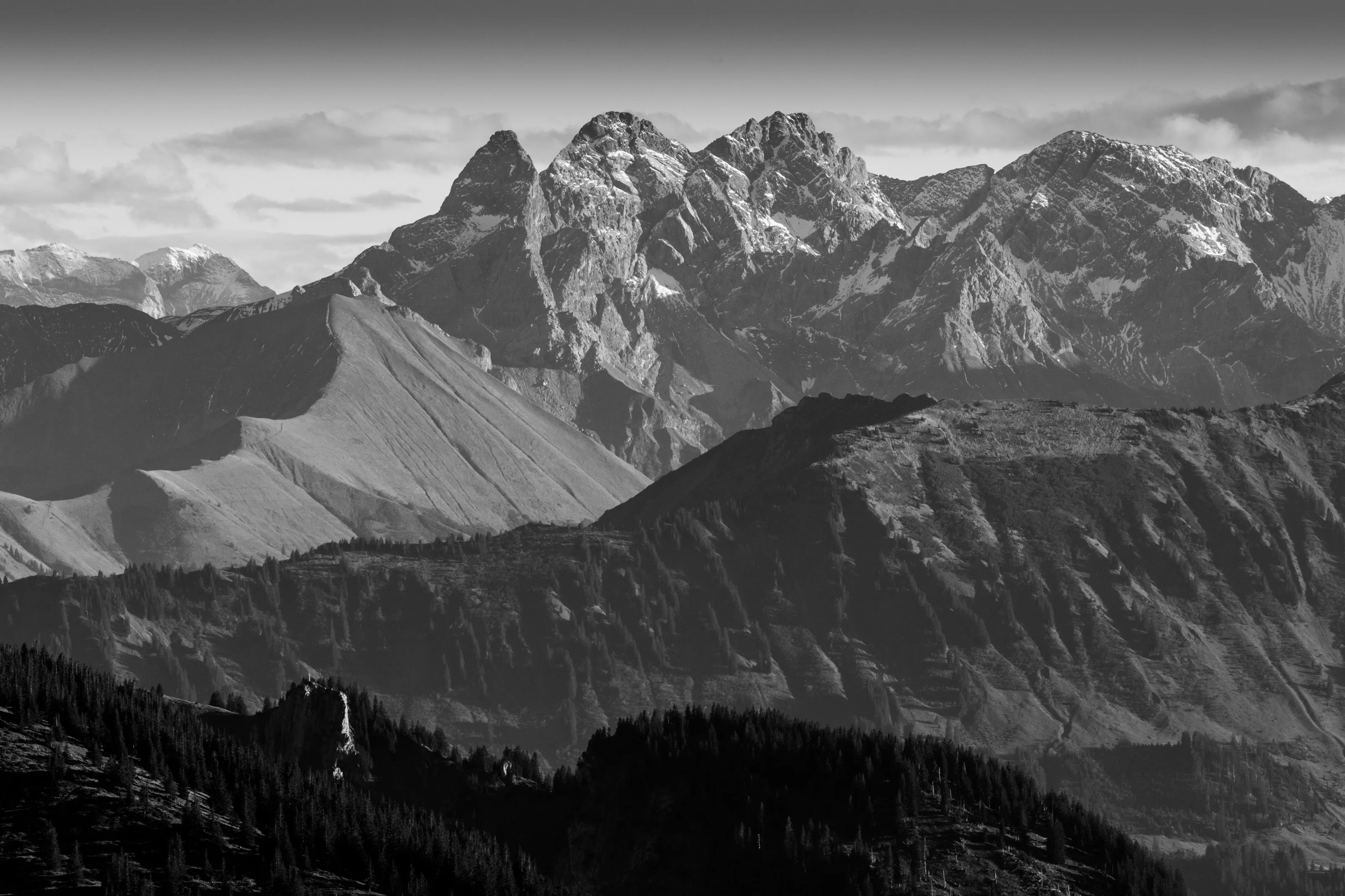 Schwarzweißaufnahme eines zerklüfteten Bergmassivs über Wald.