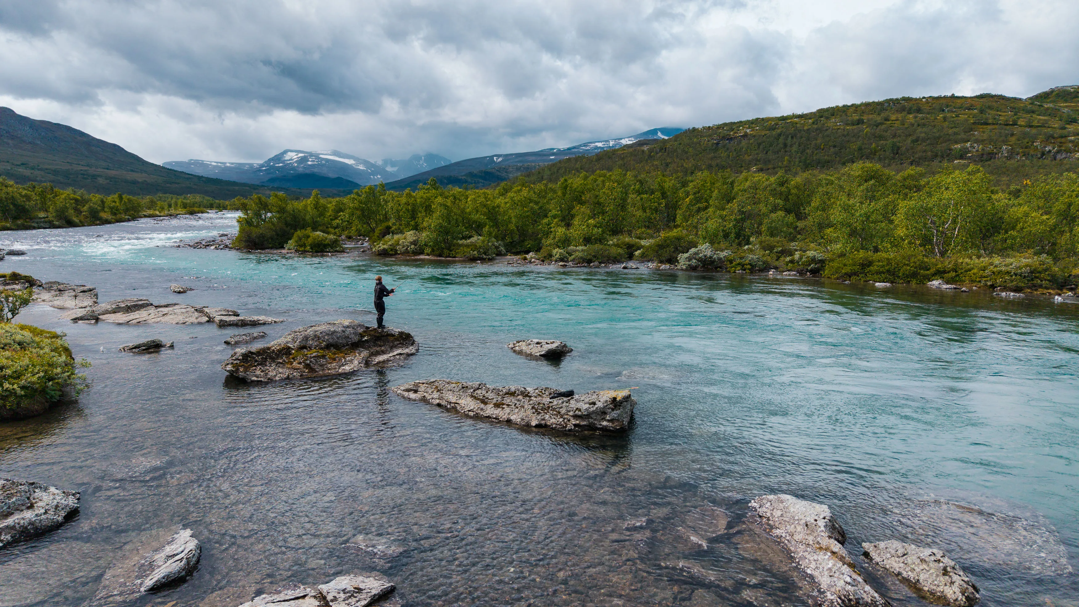 Angler steht auf einem Felsen in einem türkisfarbenen Fluss vor bewaldeten Berghängen