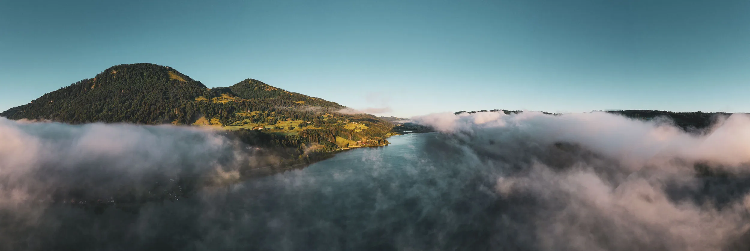 Panorama des Alpsees bei Immenstadt mit Nebel über dem Wasser und herbstlichen Hängen.