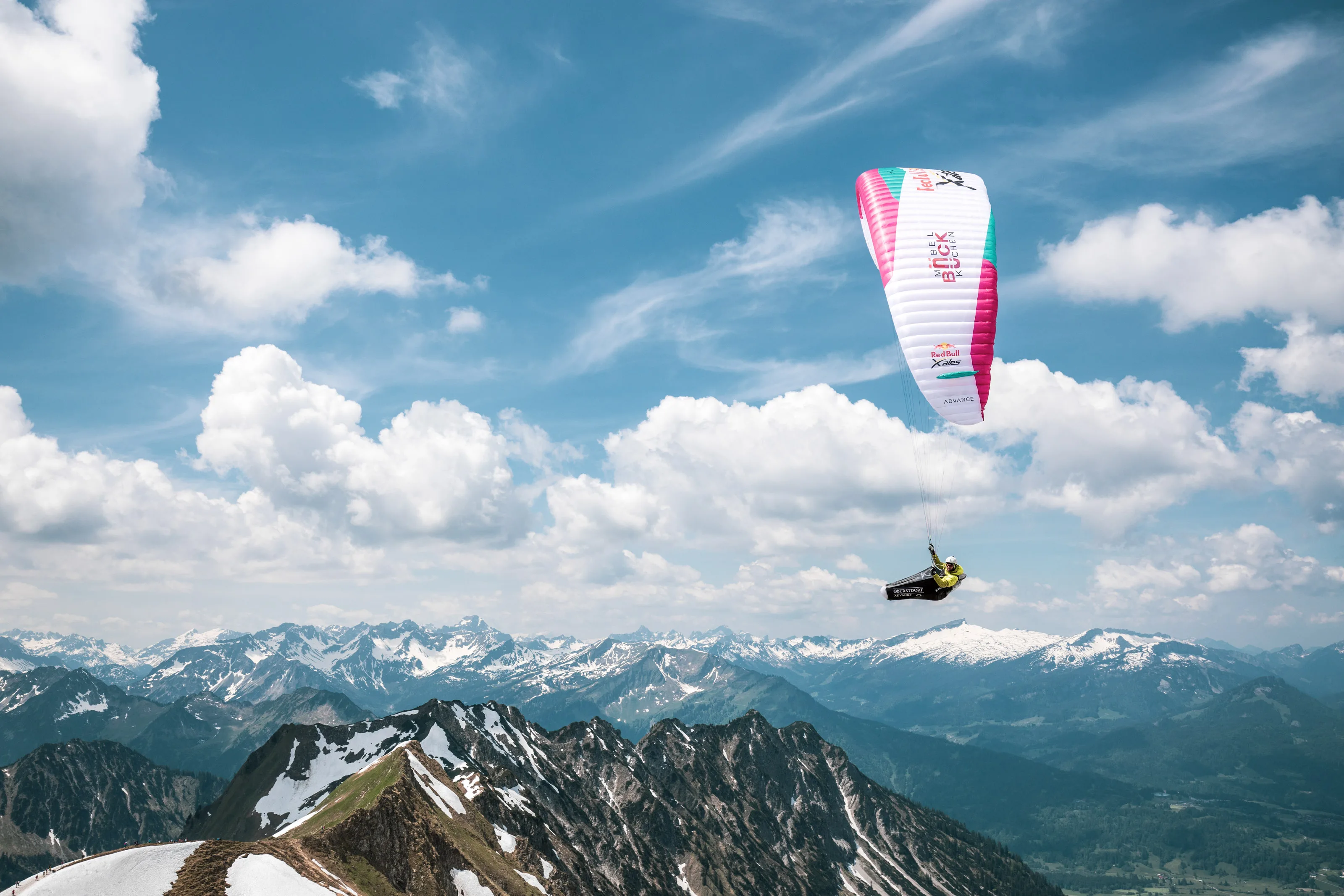 Michael Lacher fliegt unter einem pinkweißen Gleitschirm vor schneebedeckten Gipfeln.