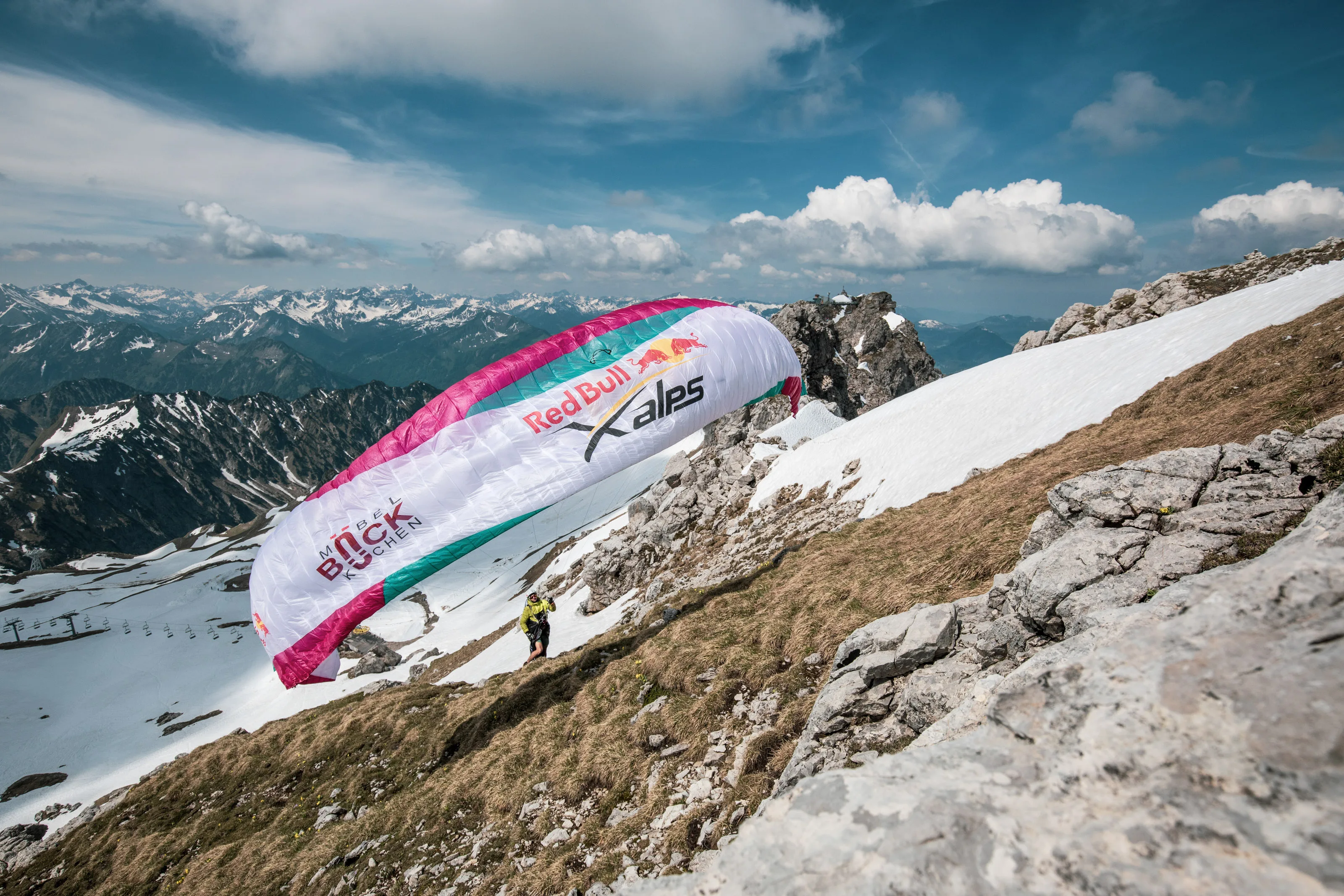 Am Hang steigt ein pinkweißer X-Alps-Gleitschirm über Michael Lacher auf; dahinter liegen verschneite Gipfel.