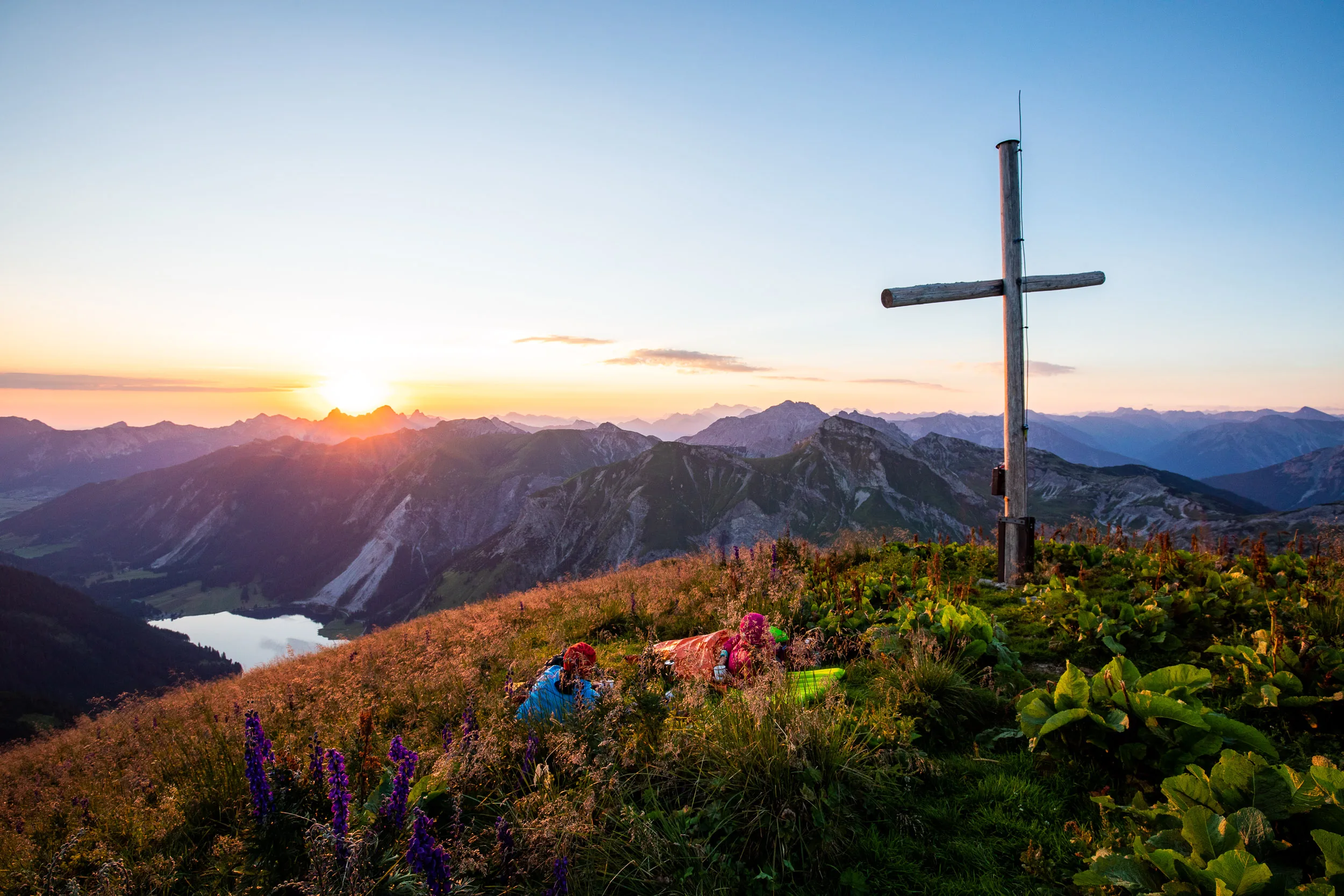 Ein Gipfelkreuz steht über einer blühenden Bergwiese vor der untergehenden Sonne