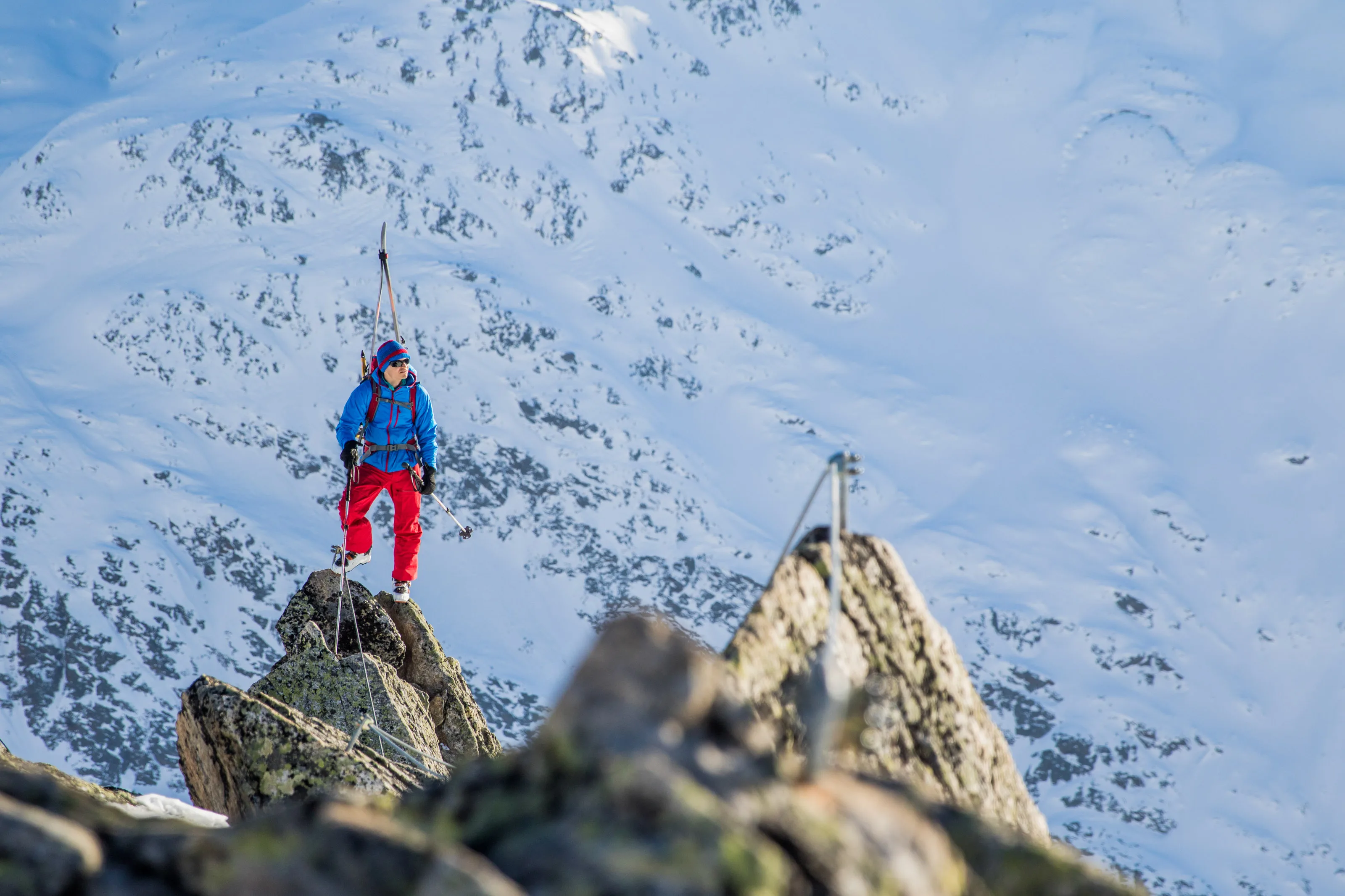 Ein Skitourengeher steht mit Tourenski auf einem Felszacken über dem verschneiten Tal