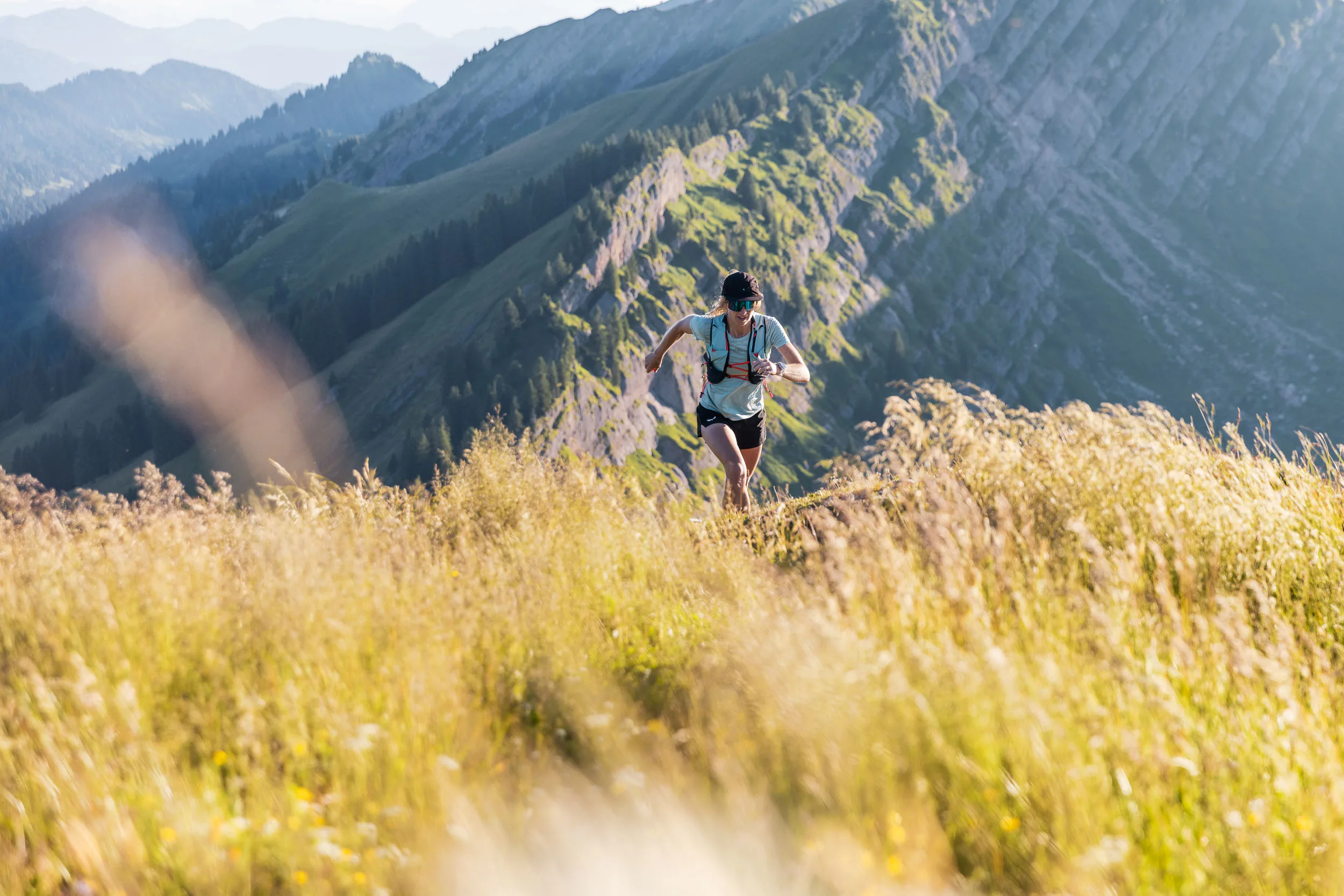 Eine Trailrunnerin läuft durch hohes Berggras auf eine Kuppe zu, hinter ihr ragen felsige Grate auf.