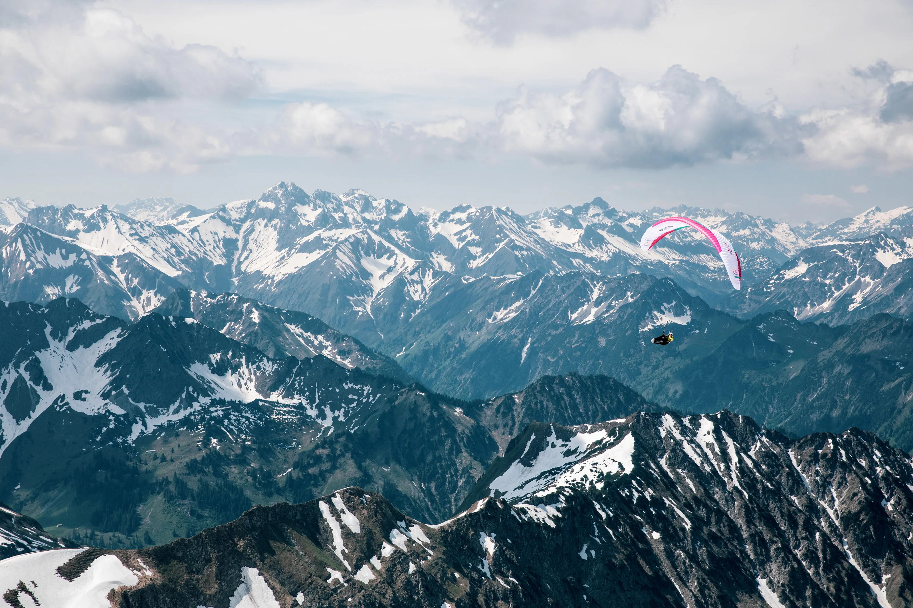 Ein pinkweißer Gleitschirm mit Michael Lacher fliegt vor einem weiten Panorama verschneiter Gipfel.
